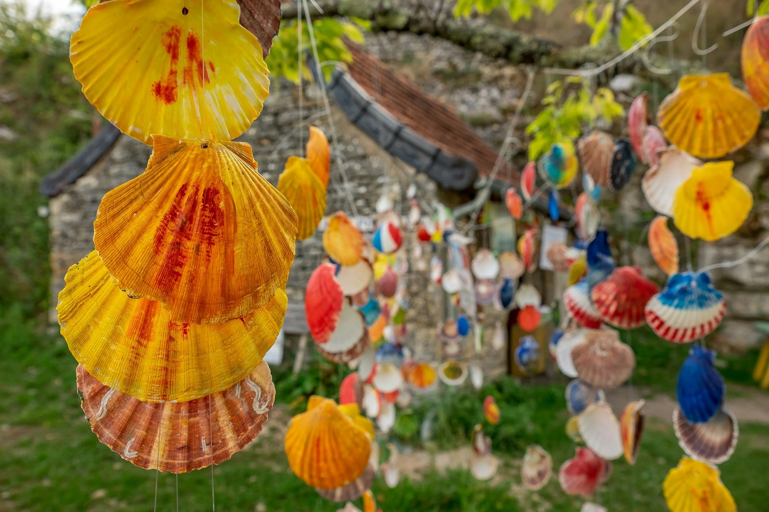 Colorful seashell hanging art installation outdoors with a rustic stone building and greenery in the background.