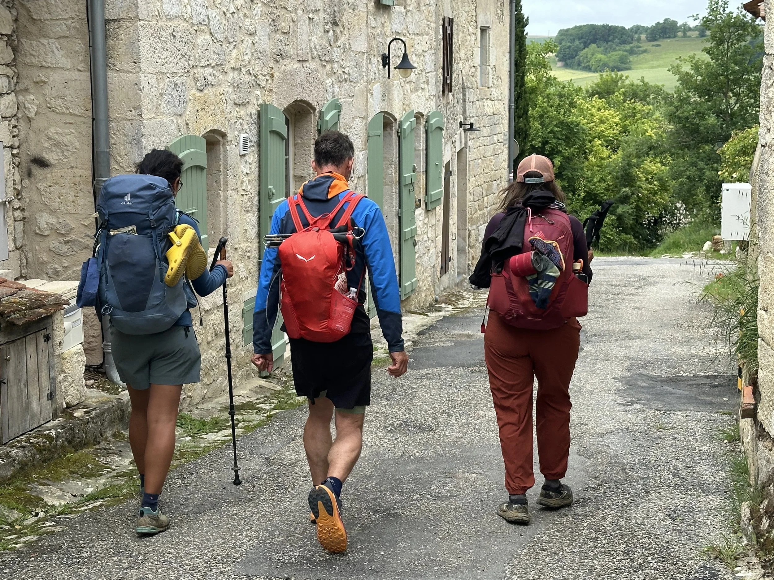 Three hikers walking down a village street with stone buildings and green trees in the background.