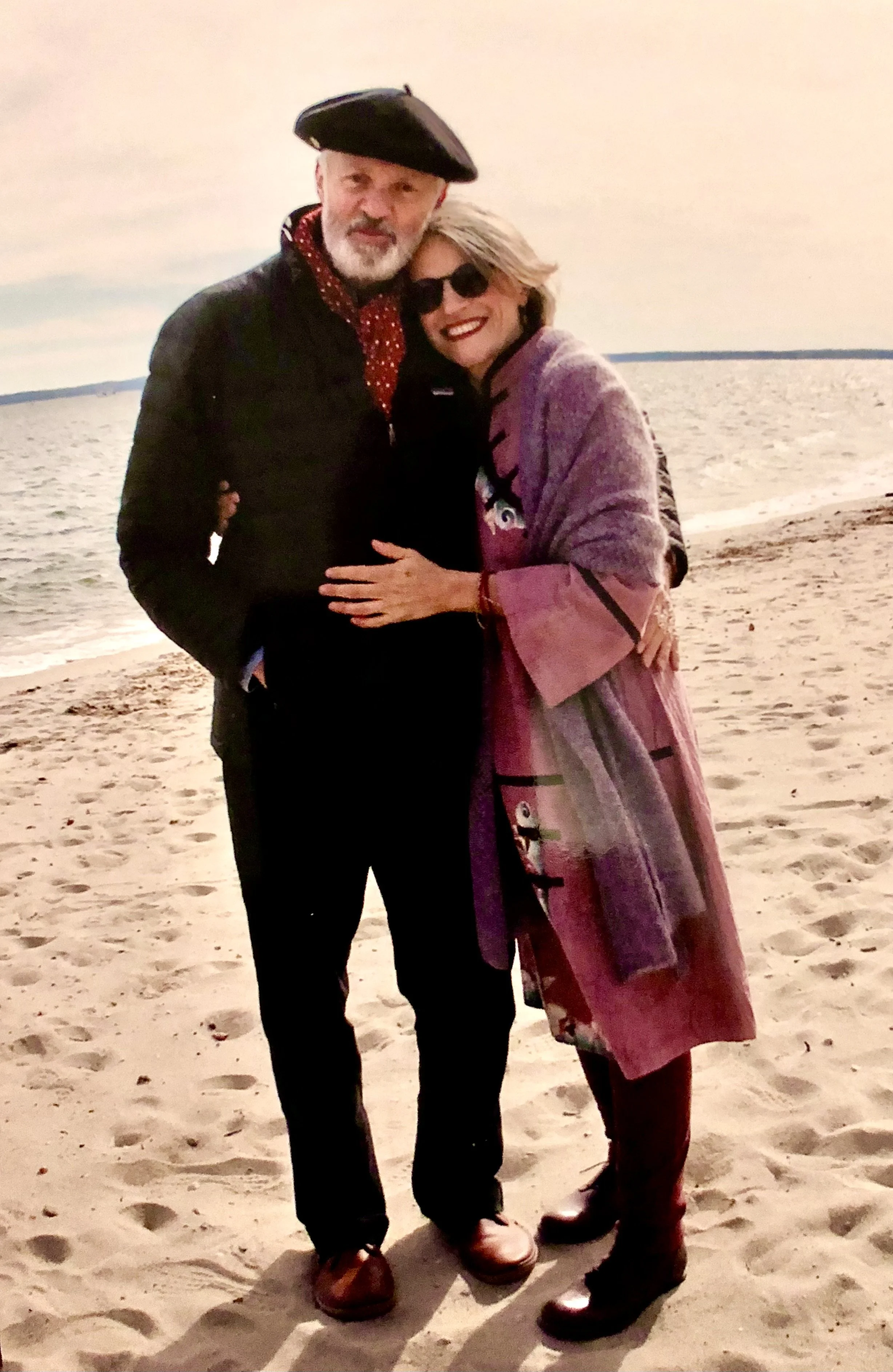 A man and woman standing close together on a sandy beach, with the sea and sky in the background. The man has a beard and is wearing a black beret, black jacket, and brown shoes. The woman is wearing sunglasses, a colorful dress, a long pink and purple coat, and brown boots. They are smiling and embracing each other.