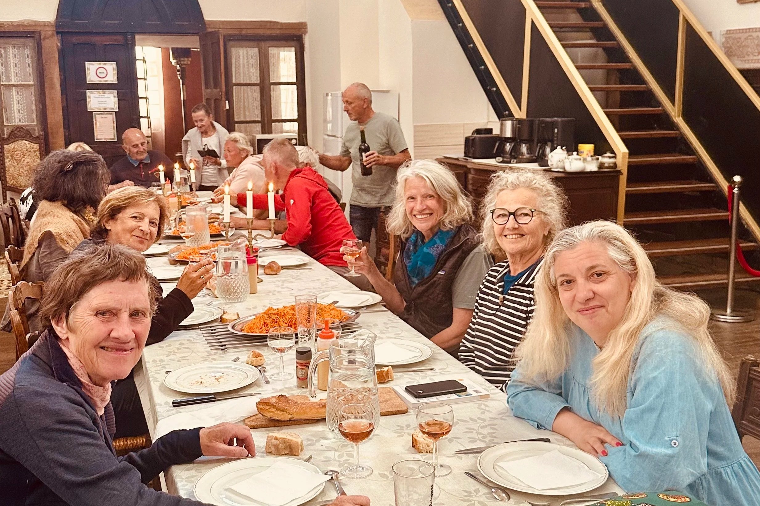 A group of adults sitting around a long dining table with food, drinks, and candles, celebrating in a warmly decorated room.