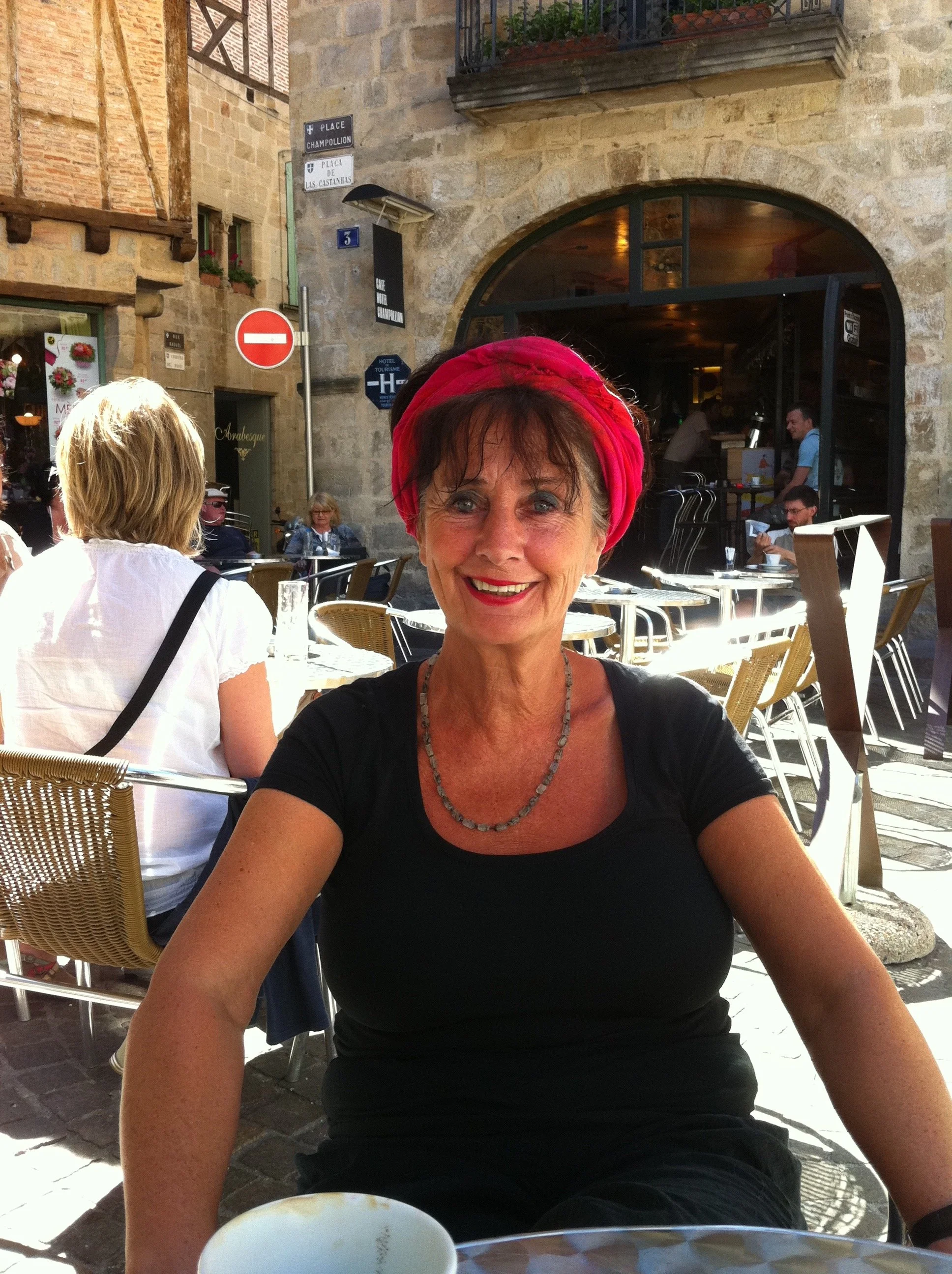 A smiling woman with a pink headscarf and black shirt sitting outdoors at a cafe in a European city.