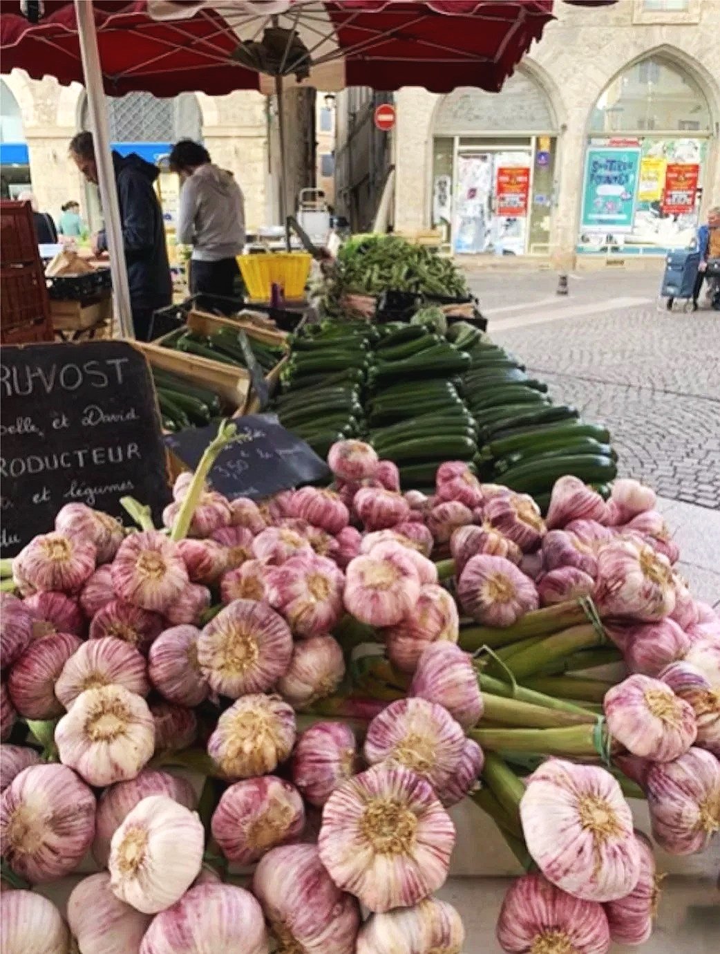 Fresh garlic and zucchinis for sale at an outdoor market stall, with two people shopping in the background and umbrellas overhead.