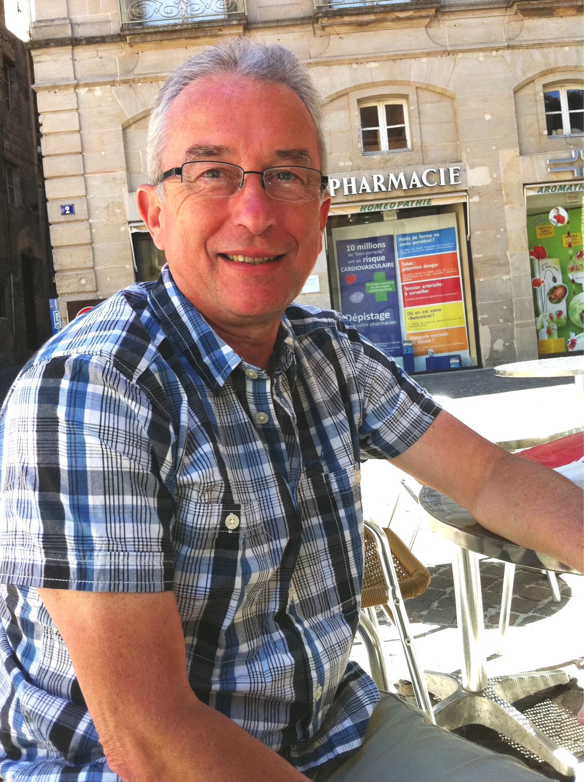 A smiling man with gray hair, glasses, and a checkered shirt sitting at an outdoor table near a pharmacy on a city street.