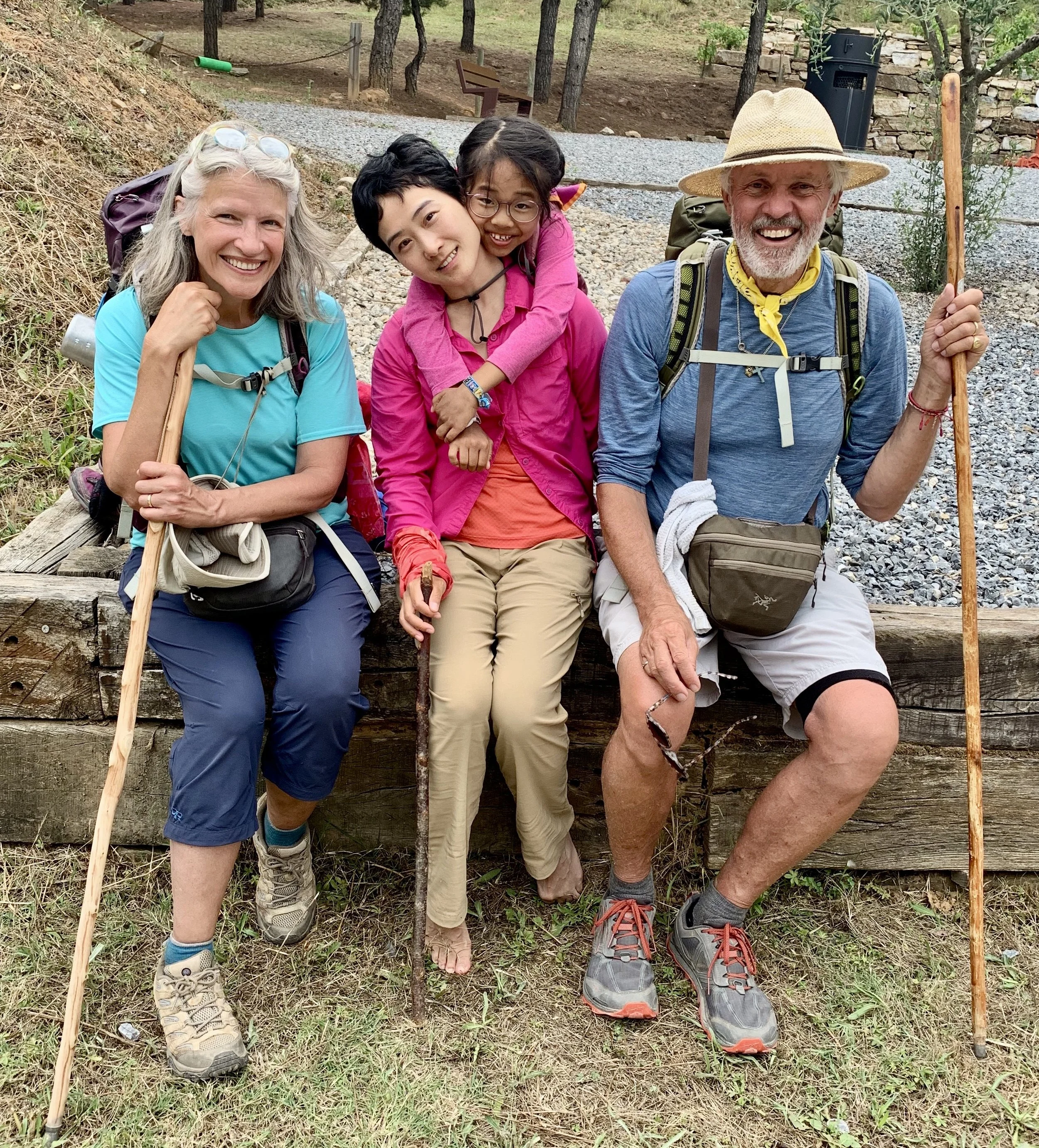 A group of four smiling hikers, two women and two children, sitting on a wooden ledge in a wooded outdoor setting. They are dressed in outdoor gear, holding hiking sticks, and appear happy after a hike.