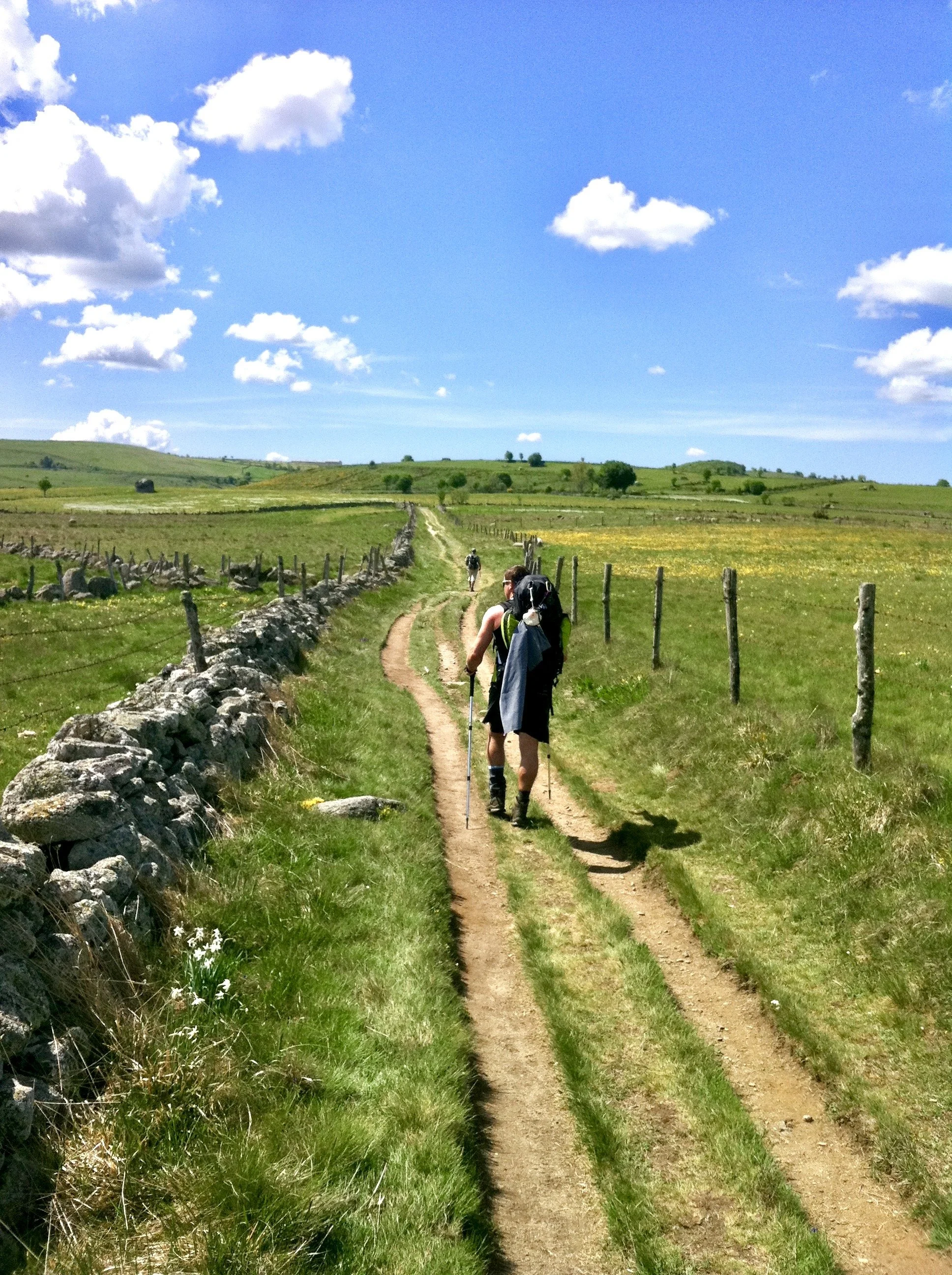 Hikers walking on a dirt trail through green countryside under a bright blue sky with white clouds.