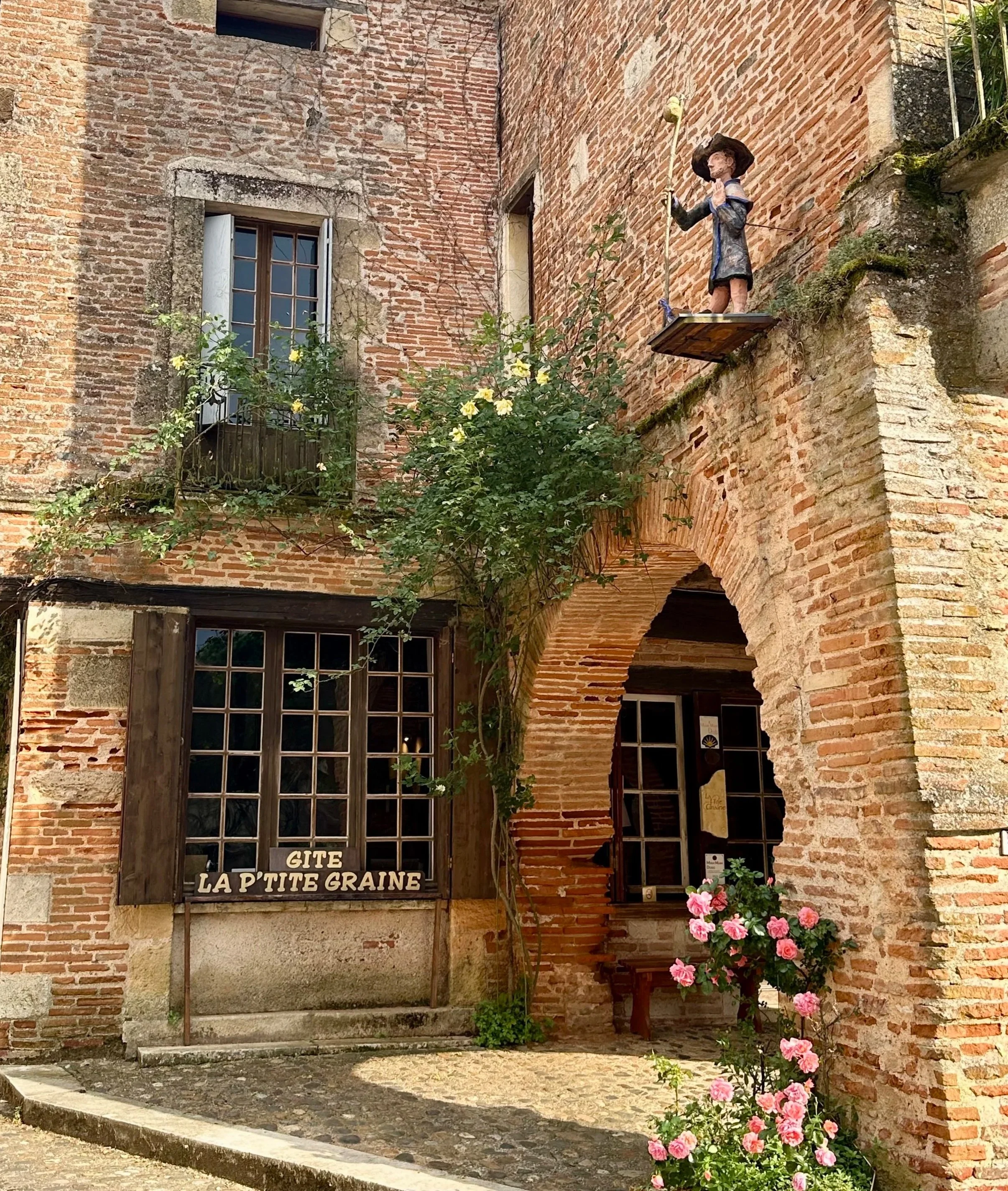 A brick building with a narrow arched doorway, a window with wooden shutters, and a small balcony with open shutters. There is a decorative figurine of a girl with a hat and a walking stick on a ledge, surrounded by greenery and pink flowers outside.