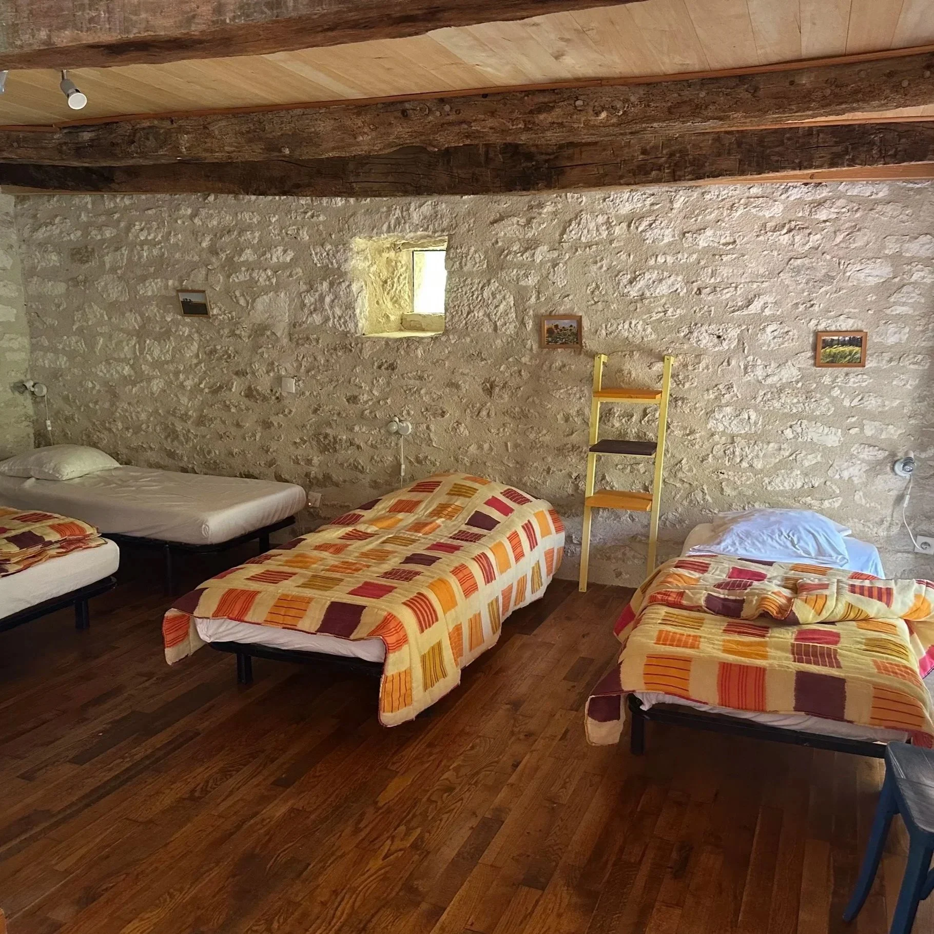 A rustic bedroom with three single beds, each with colorful striped bedding. Two beds have pillows, and one bed has a blanket. The room has a stone wall with a small window, and a wooden beam ceiling. There are a few small pictures on the wall and a yellow ladder shelf between two beds. The floor is wood.
