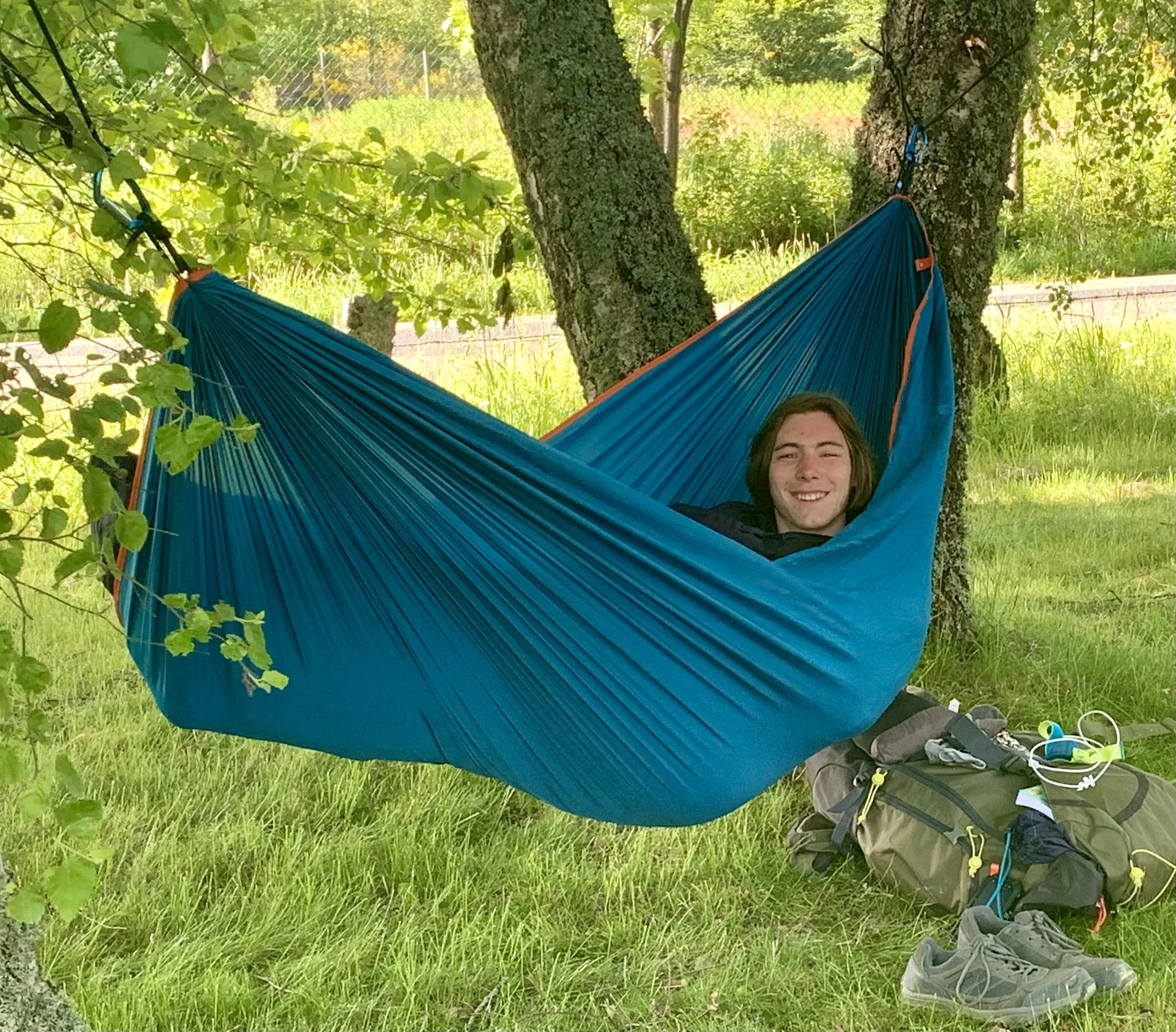 A person lying in a blue hammock tied between two trees in a grassy outdoor setting.