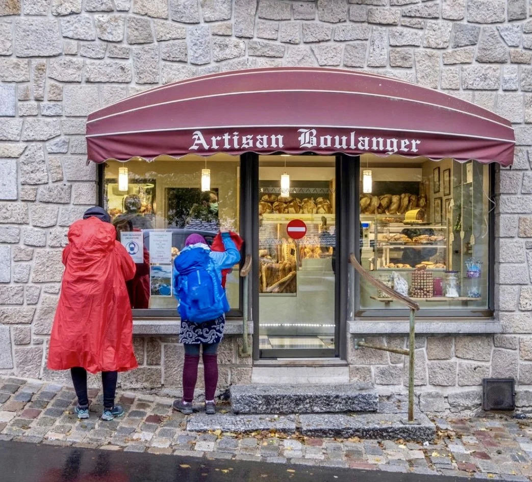 People standing outside a bakery with a maroon awning that reads "Artisan Boulanger". The bakery has a window display of bread and pastries, and a "do not enter" sign on the door.