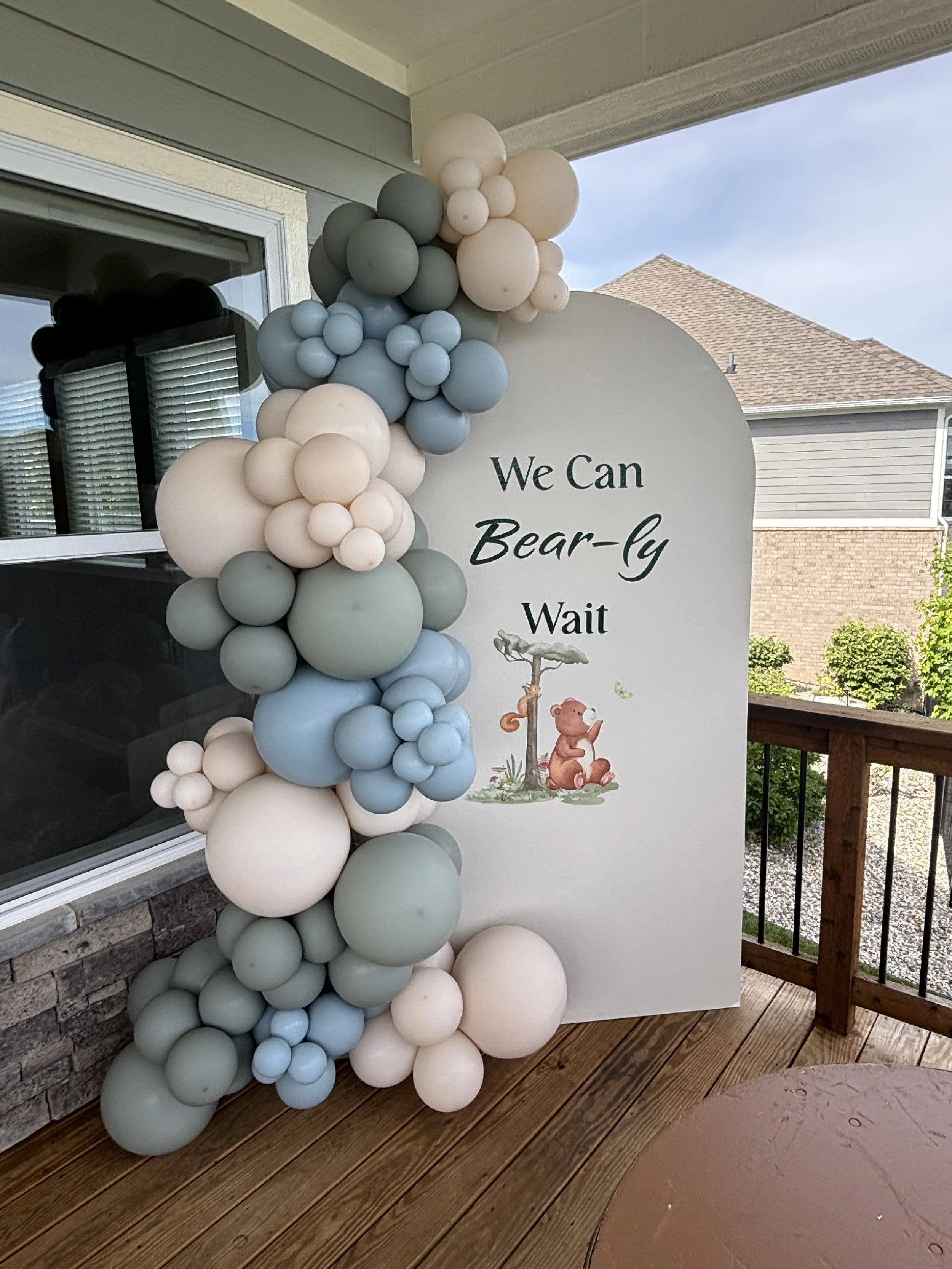 Decorative arch of white, gray, and light blue balloons next to a sign with text and a bear illustration, on a wooden deck outside a house.