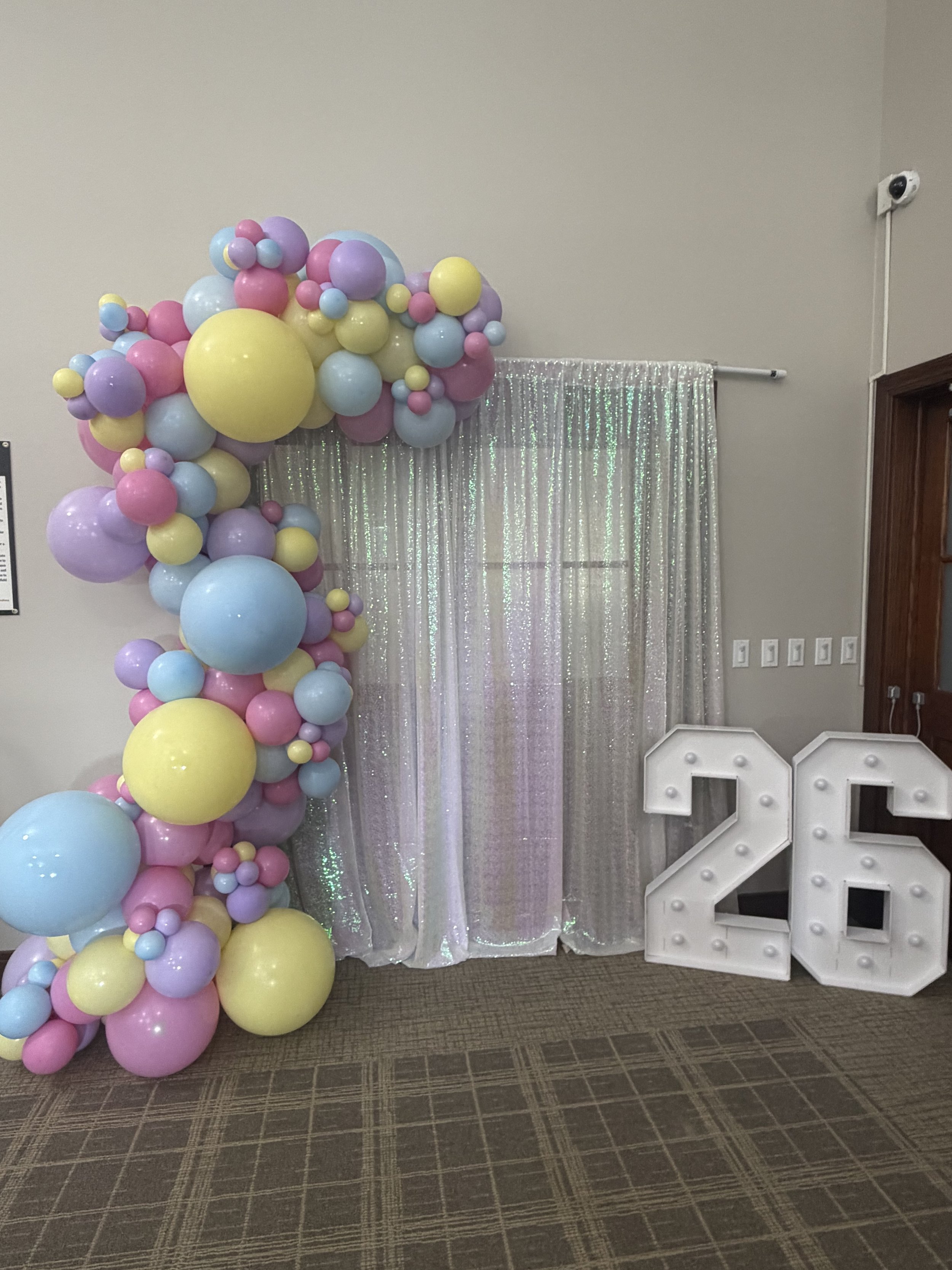 Decorative display with pastel colored balloons forming a vertical arrangement next to a silver sequin curtain, with a large illuminated '2024' sign on the right.