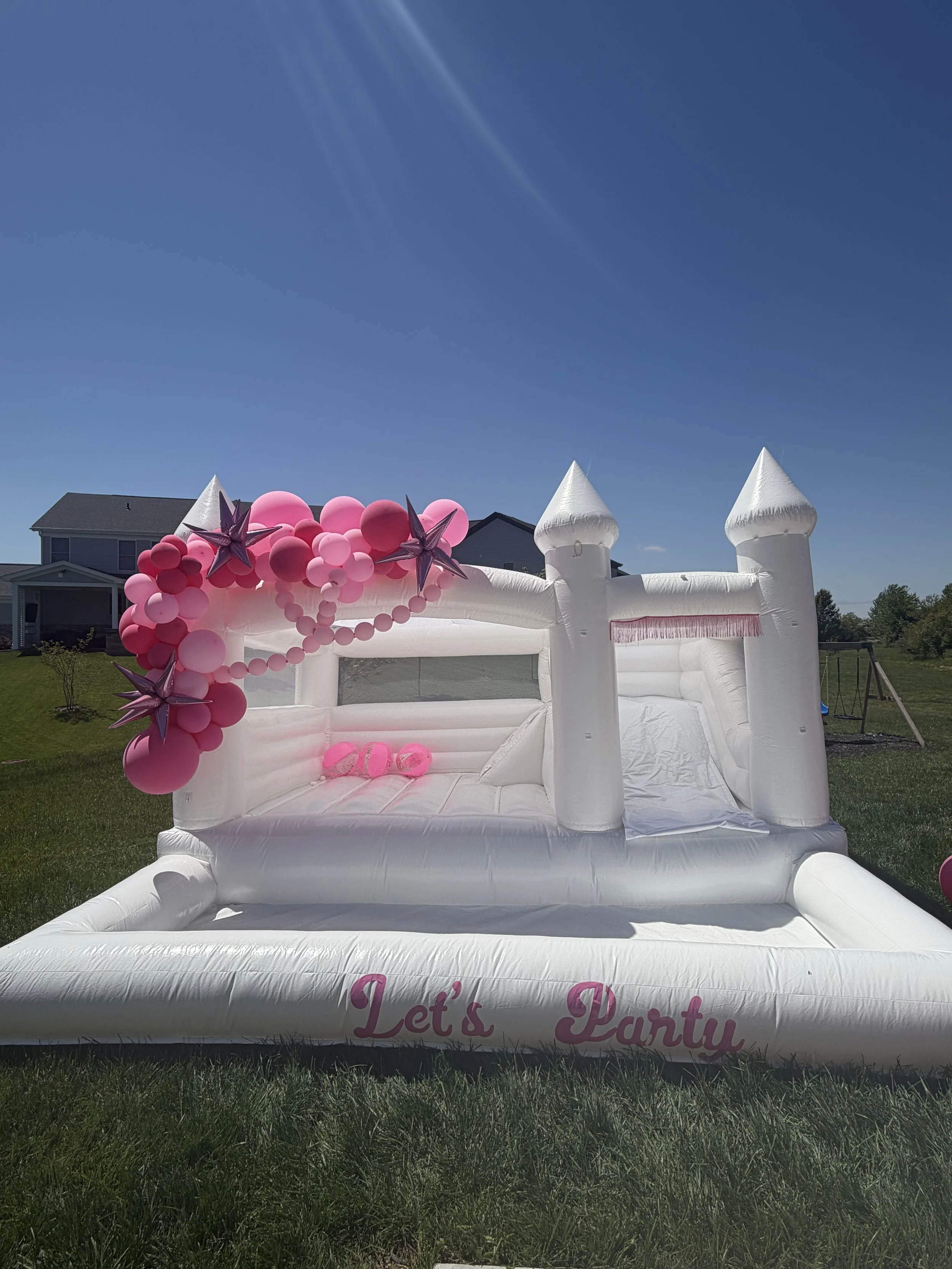 A white inflatable bouncy house with turrets, decorated with pink balloons and purple star-shaped balloons, set outdoors on grass with houses in the background, under a clear blue sky.