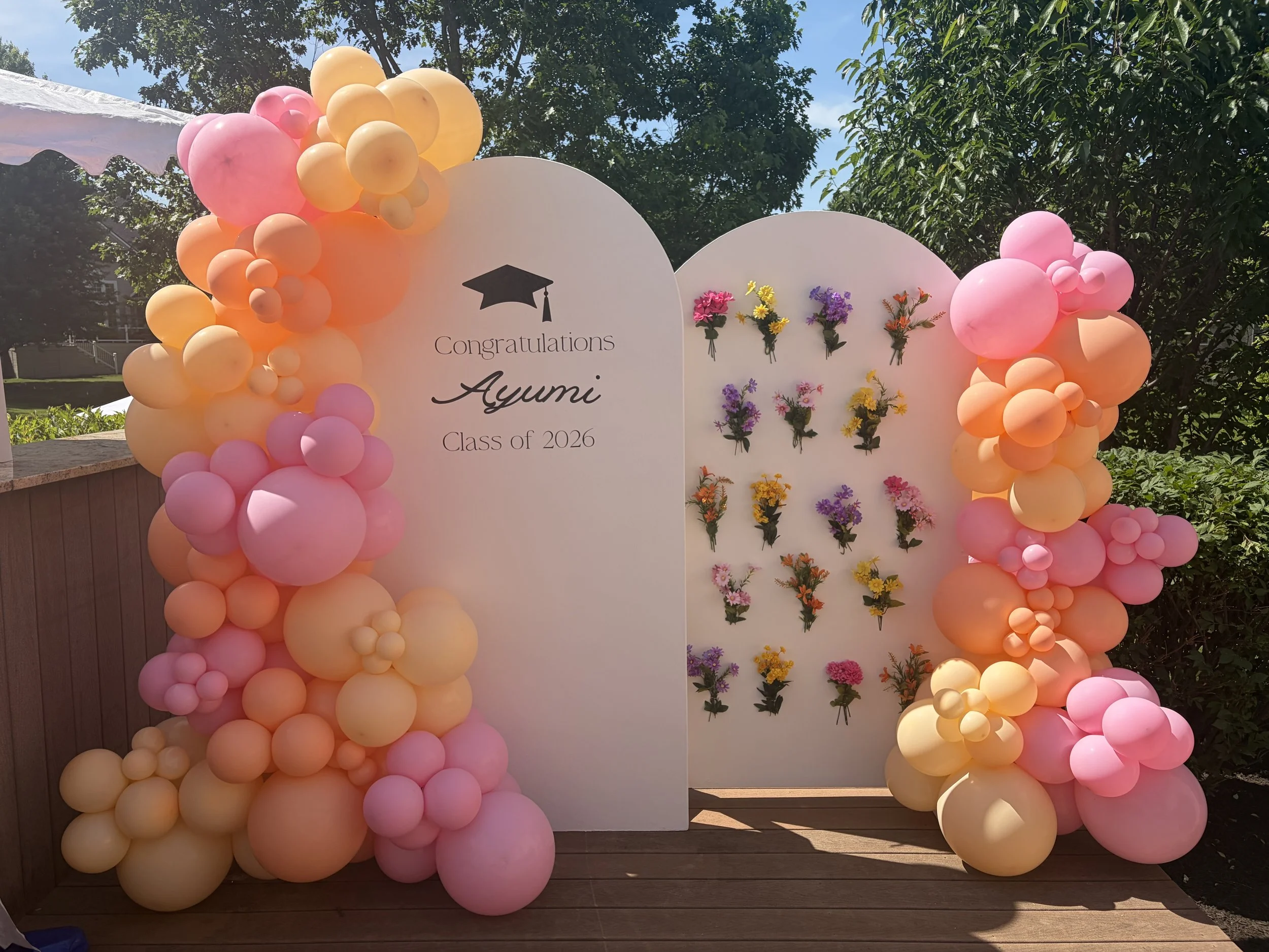 Decorative backdrop for a graduation celebration with pink, peach, and yellow balloons, floral arrangements, and a sign that says "Congratulations Ayumi Class of 2026."