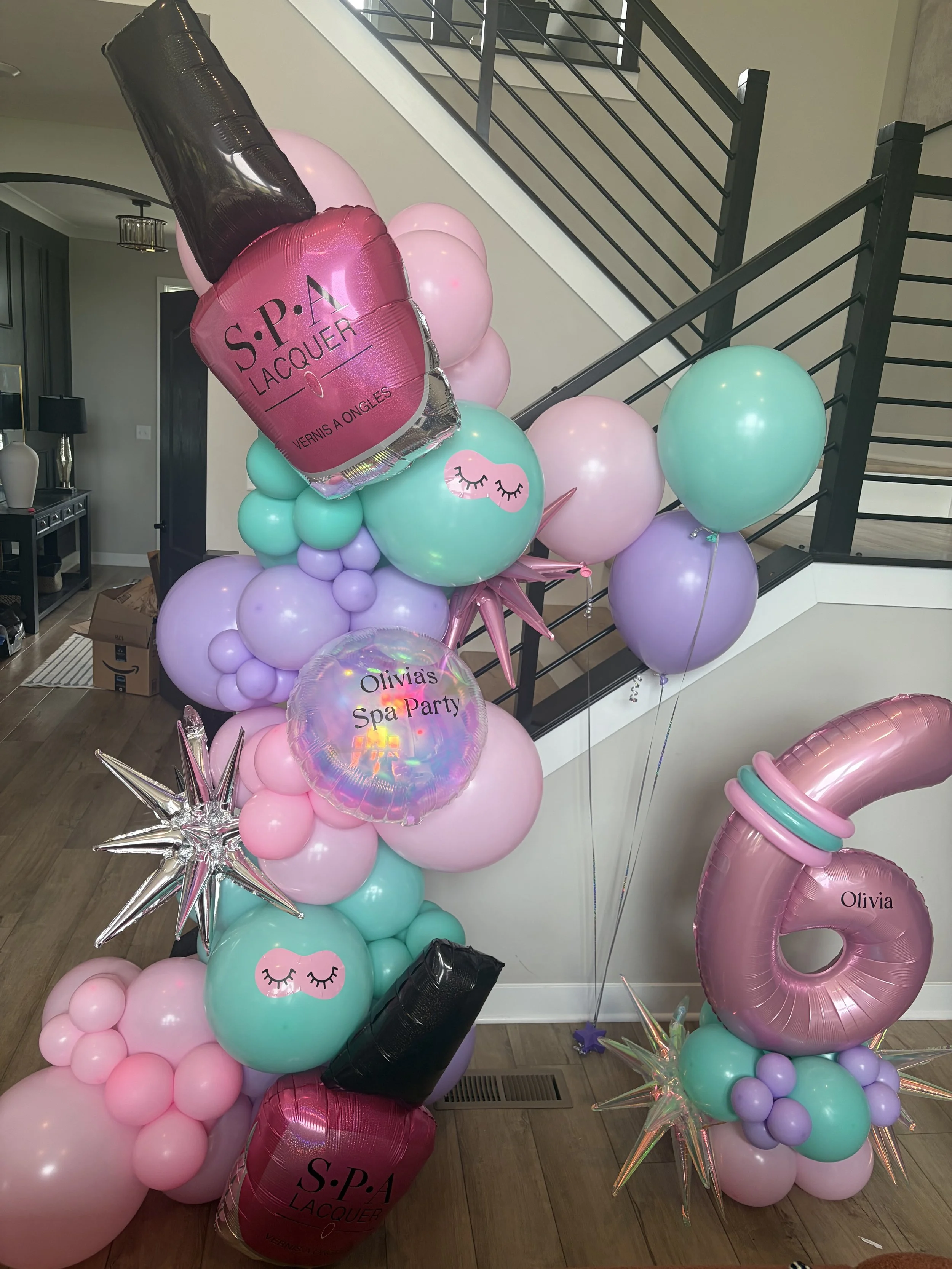 Colorful balloon arrangement for a spa party, including pink, mint, purple, and black balloons, some with eyelash designs, star-shaped balloons, and one with the text 'Olivia's Spa Party'.