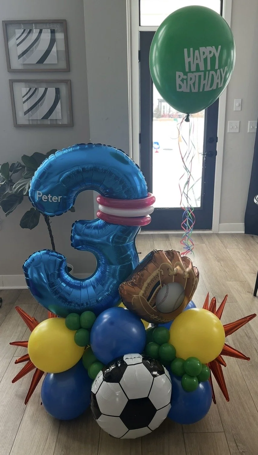 Balloon arrangement with a large blue number 3, a green balloon with "Happy Birthday," a soccer ball balloon, a baseball mitt holding a baseball balloon, and various green and blue balloons.