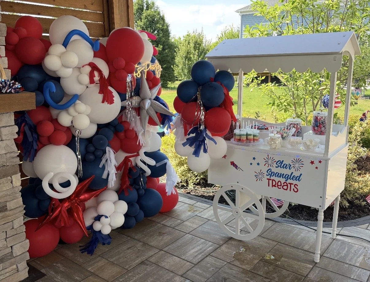 Patriotic-themed balloon decoration with a white candy cart labeled 'Star Spangled Treats' outside under a covered patio, with a garden and house in the background.