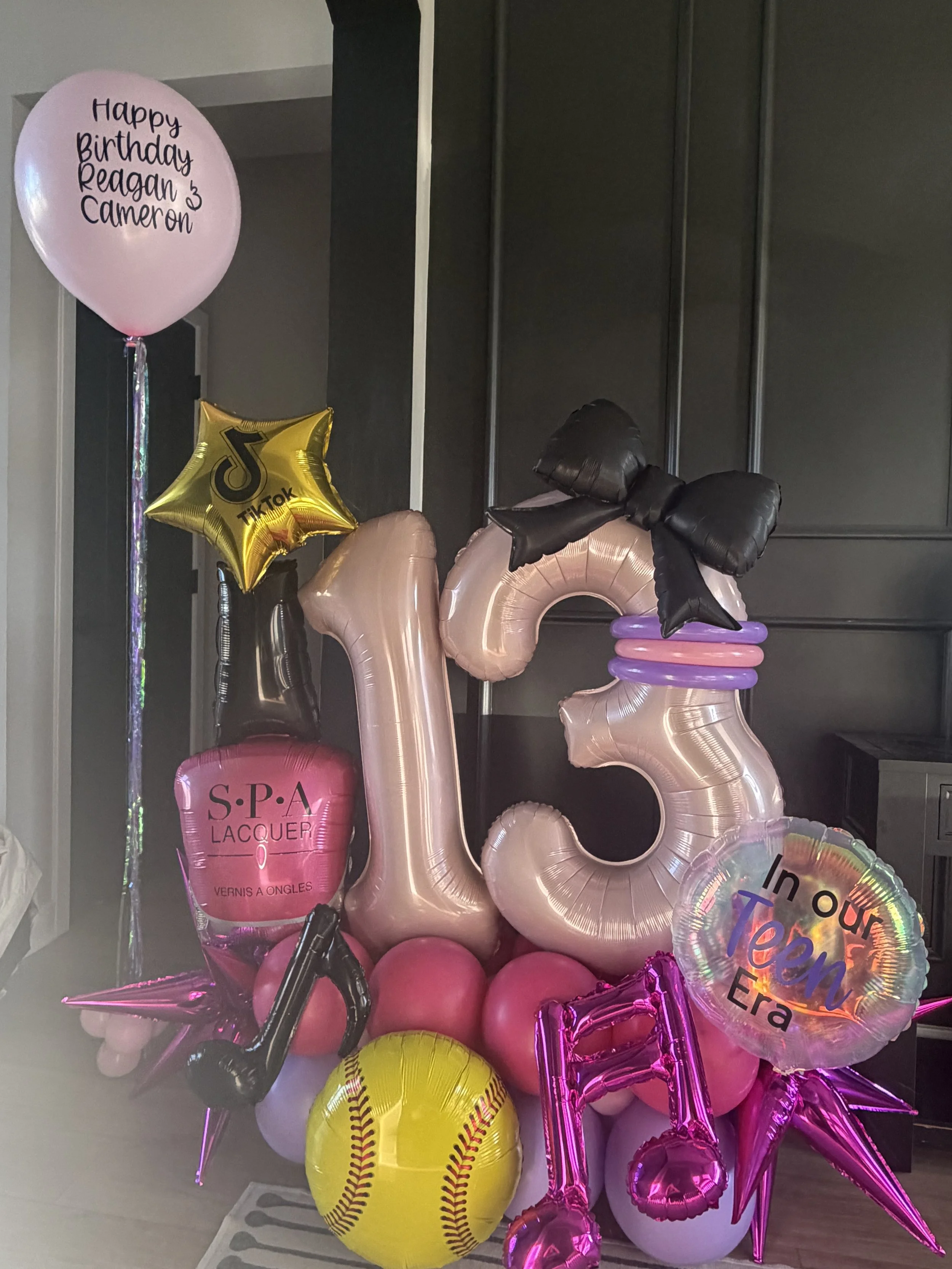 A collection of balloons including a pink balloon with the message 'Happy Birthday Reagan 3 Cameron,' a Gold TikTok star-shaped balloon, a pink number 3 balloon, a yellow baseball-shaped balloon, a pink spa balloon, and other pink and purple balloons, some with messages and others plain.