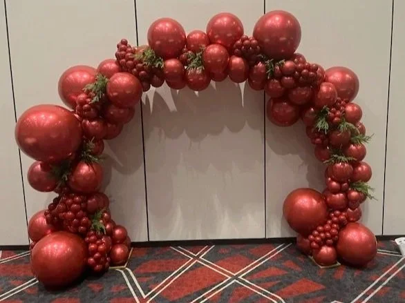 Decorative arch made of red Christmas ornaments and small green pine branches, set against a gray wall on a patterned carpet.