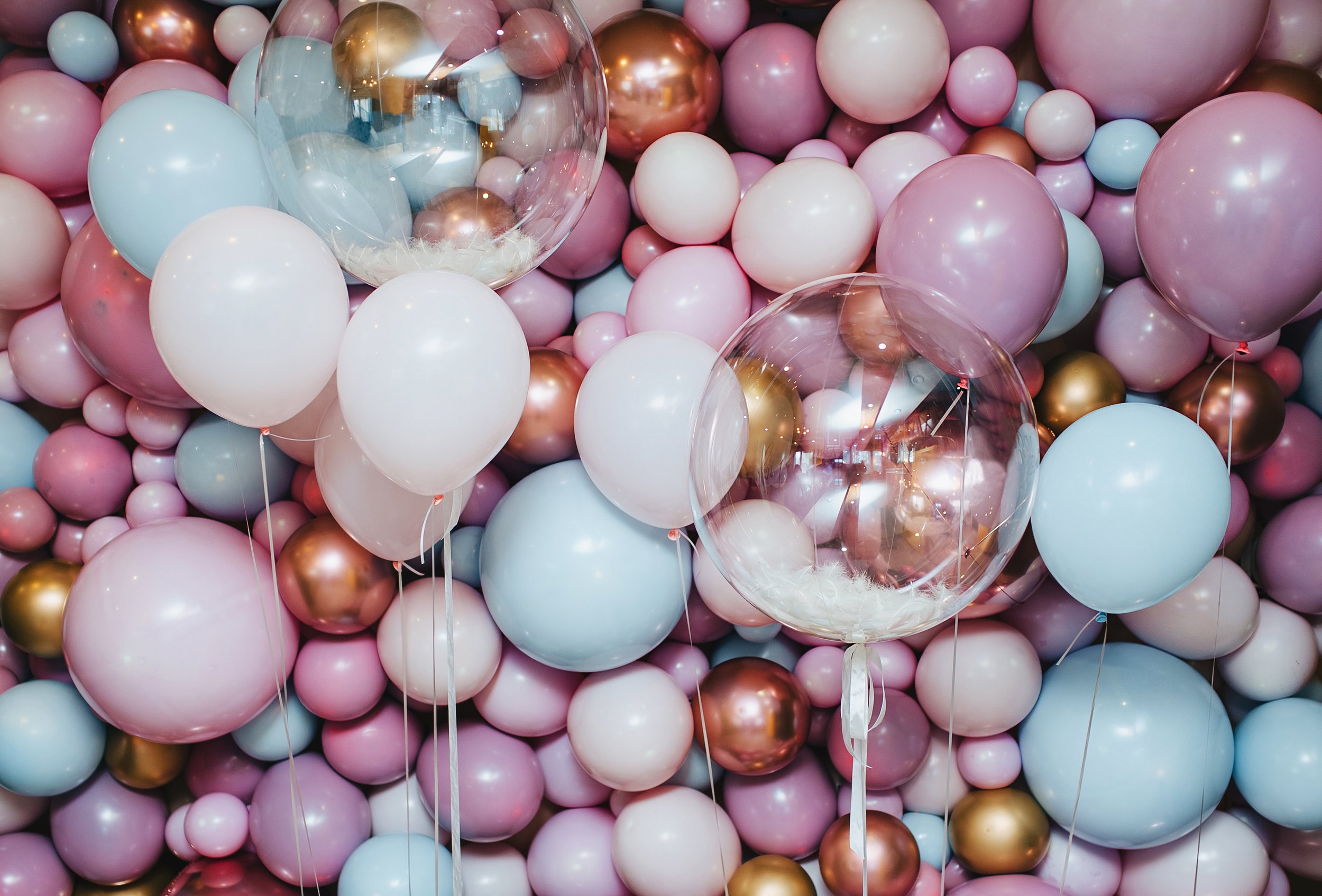 A large assortment of pastel-colored and metallic balloons, primarily pink, white, blue, and gold, filling the entire frame, with some clear balloons reflecting the surroundings.
