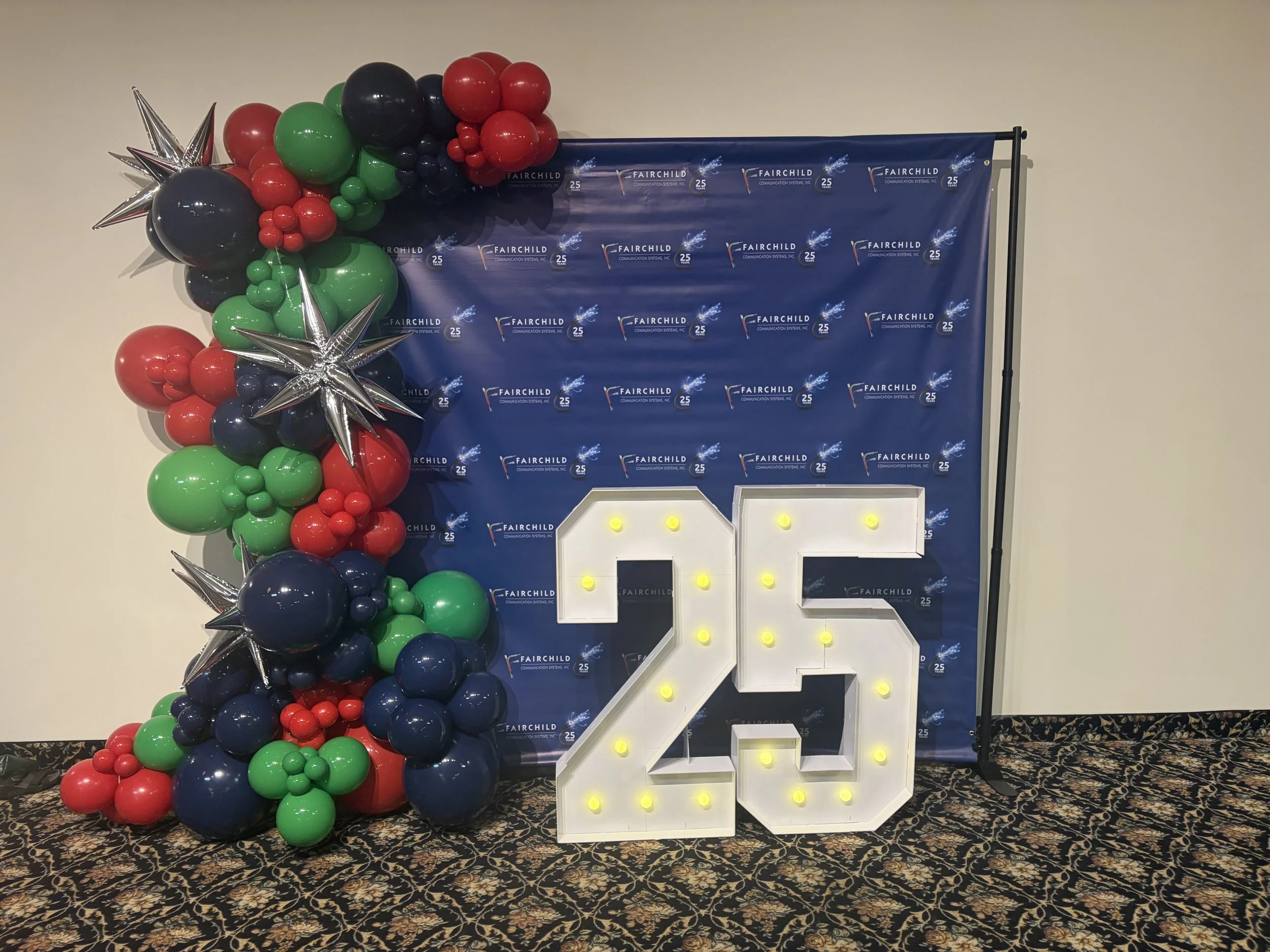 Decorative balloon arrangement featuring black, red, green, and silver balloons with silver star-shaped balloons on the left, set against a blue backdrop with the logo of Fairchild Communication Systems, Inc., and a large lit-up number 25, indicating a celebration or anniversary.