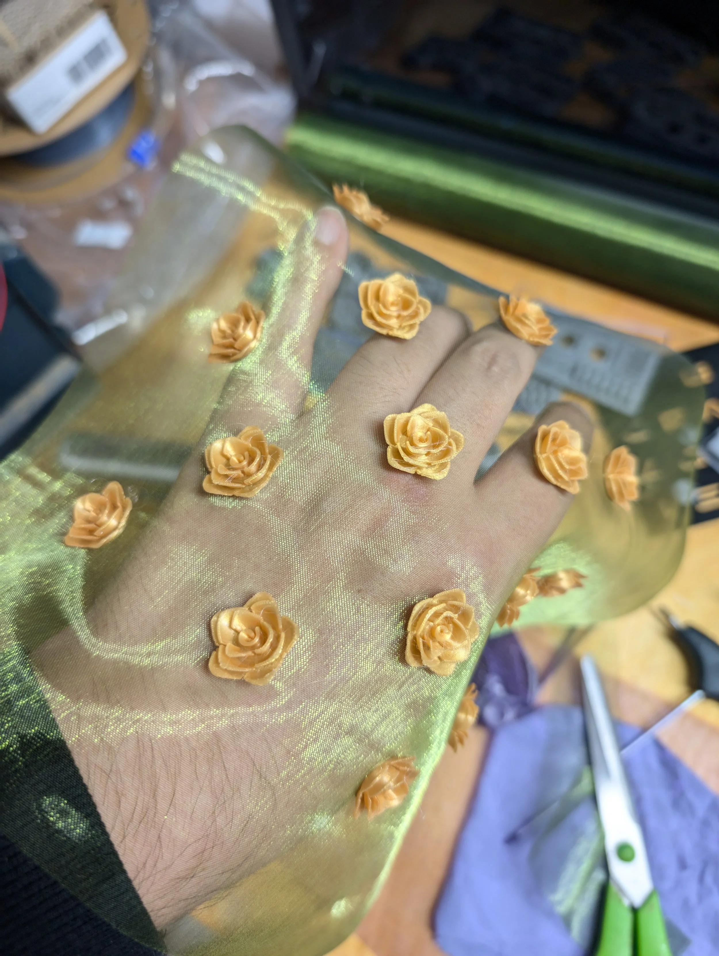 Hand trägt transparenten Stoff mit kleinen goldfarbenen Rosenapplikationen, in einem Bastelraum mit Schere, Papier und anderen Bastelmaterialien im Hintergrund.