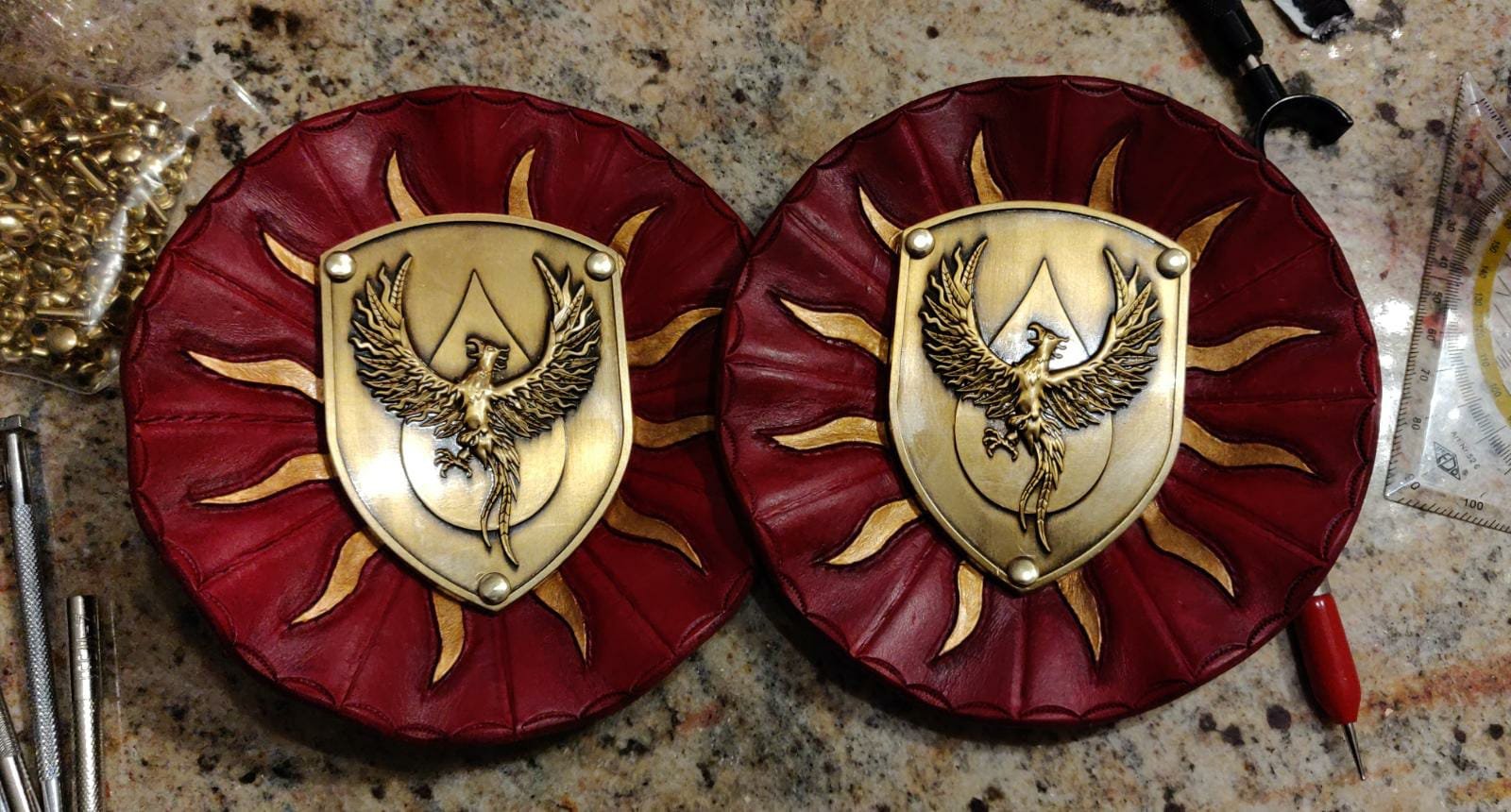 Zwei gepanzerte Schilde mit goldenen Adlerwappen auf rotem Leder auf einem Werkbank, umgeben von Werkzeugen.