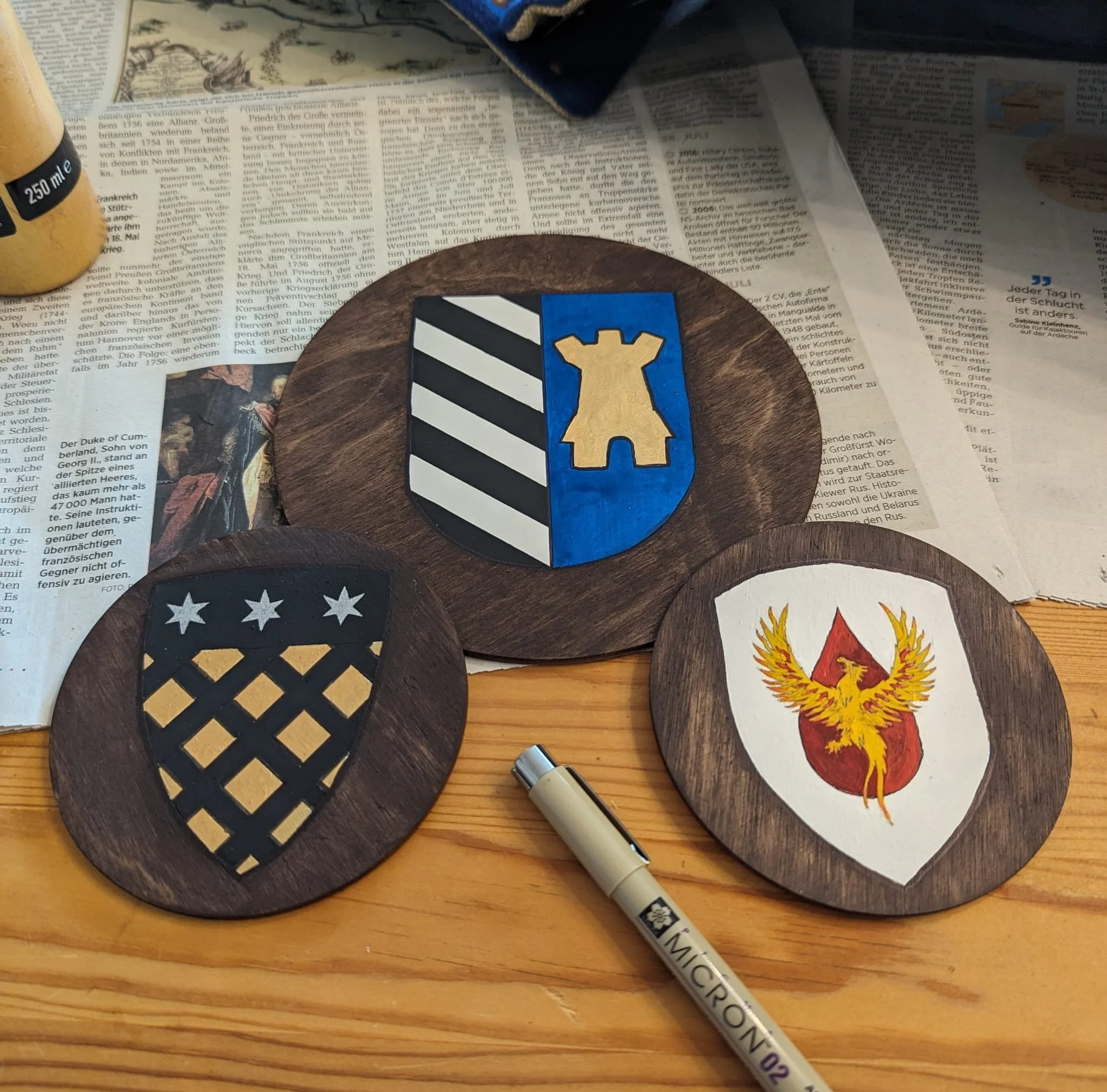 Drei Holzplatten mit Wappen oder Emblemen darauf, auf einem Tisch mit Zeitung, einem Marker und einer Flasche. Das zentrale Wappen ist blau mit einer goldenen Burg. Das linke Wappen ist schwarz mit weißen und goldenen Mustern, das rechte Wappen ist w