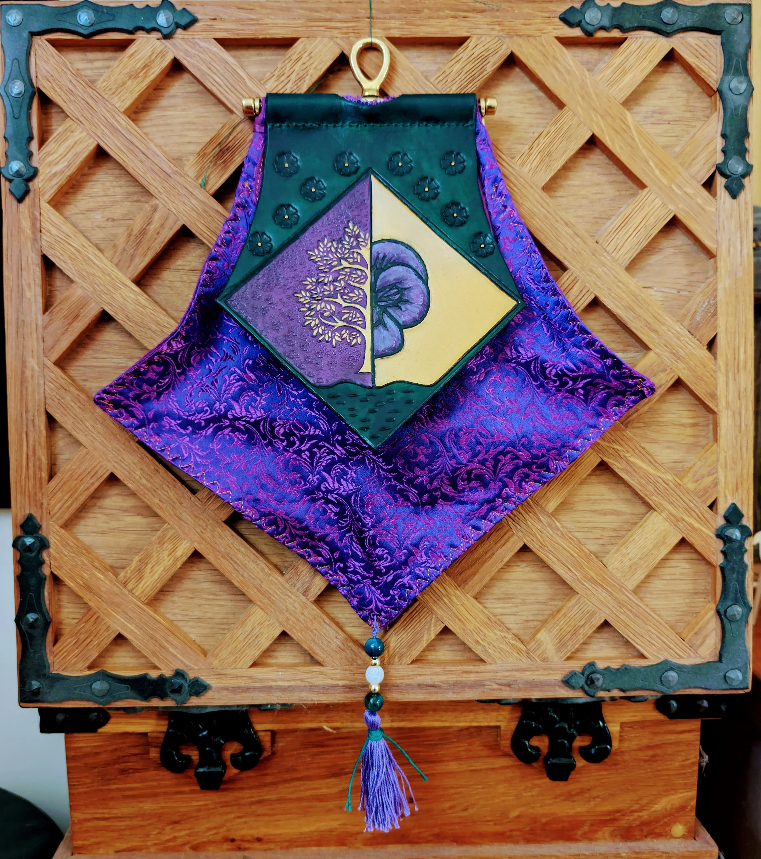 Ein kunstvoll gestaltetes Wandtuch mit einer quadratischen, bunten Stickerei, die ein Baum und eine Muschel zeigt, umrahmt von violettem und grünen Stoff, hängt an einem Holzrahmen.