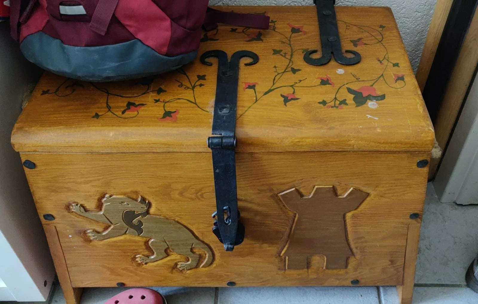 ein Holzkasten mit dekorativen Löchern, auf dem ein aufhängbares Schloss liegt, und auf dem die Abbildungen eines Löwen und einer Burg eingraviert sind, mit einer rote- und pinkfarbigen Tasche oben auf dem Kasten.