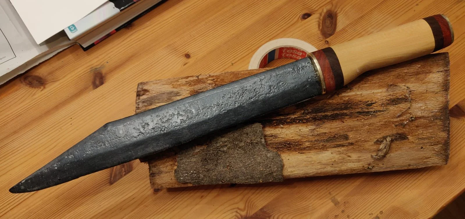 Ein mittelalterliches Messer mit einem schwarzen, unregelmäßig strukturierten Klingenstück und einem Holzgriff, das auf einem Holzblock liegt.