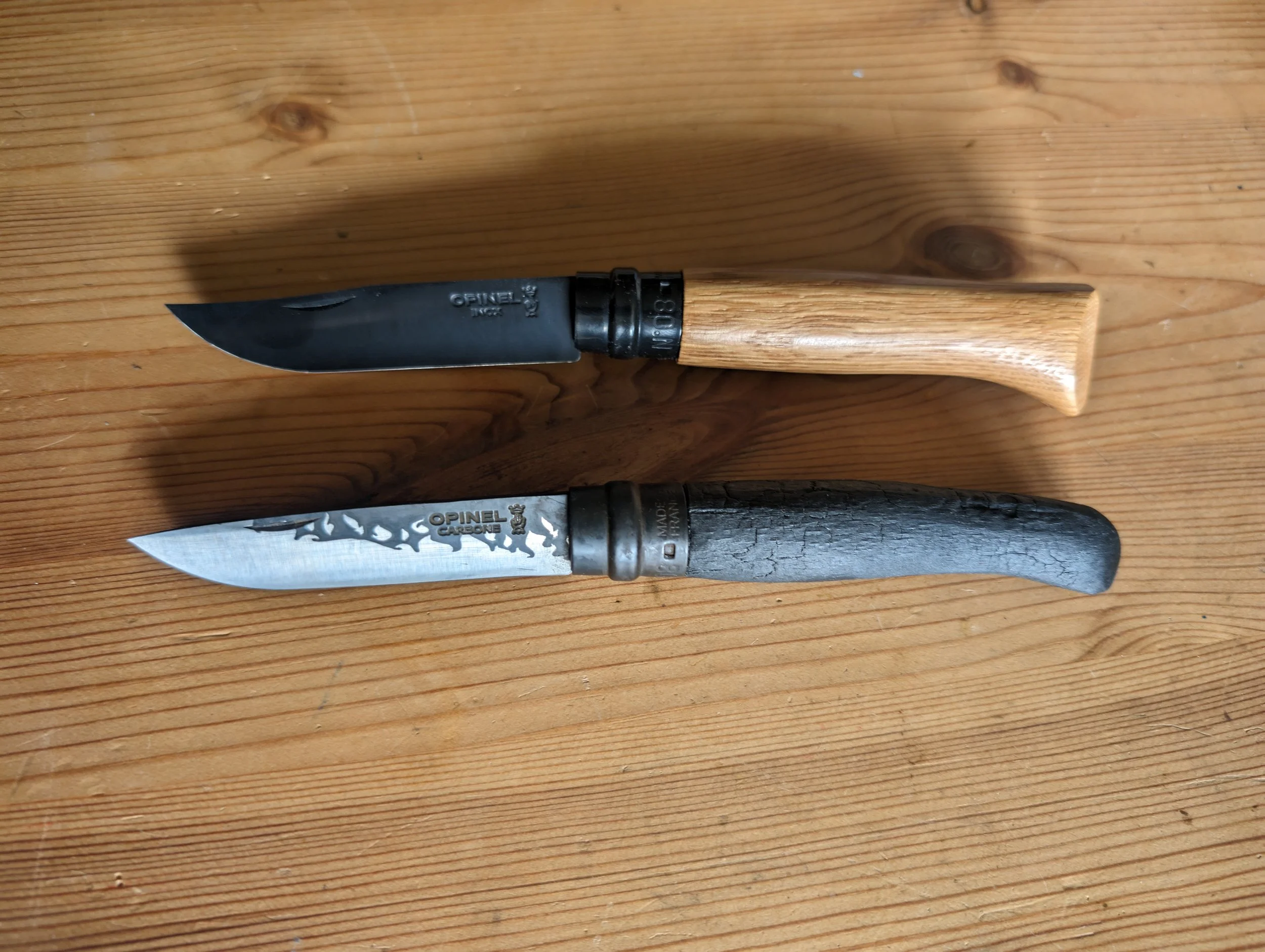 Zwei Messer mit Holz- und Griff, eines mit schwarzem Griff und einem schwarzen Messerrücken, das andere mit angewinkeltem Griff und grafischem Muster auf dem Klingenblatt, liegen auf einem Holzoberfläche.