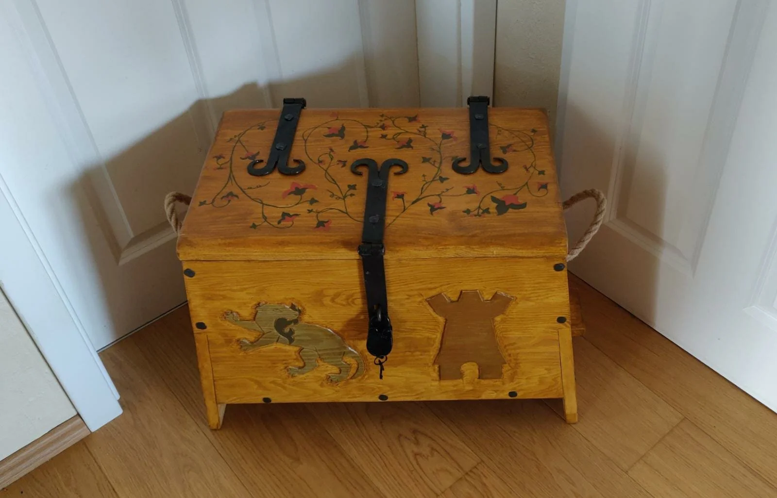 Eine bemalte Holzkiste mit Tier- und Pflanzenmotiven, stabilen Griffen und metallischen Beschlägen, steht in einer Ecke, vor einer weißen Tür, auf einem Holzboden.