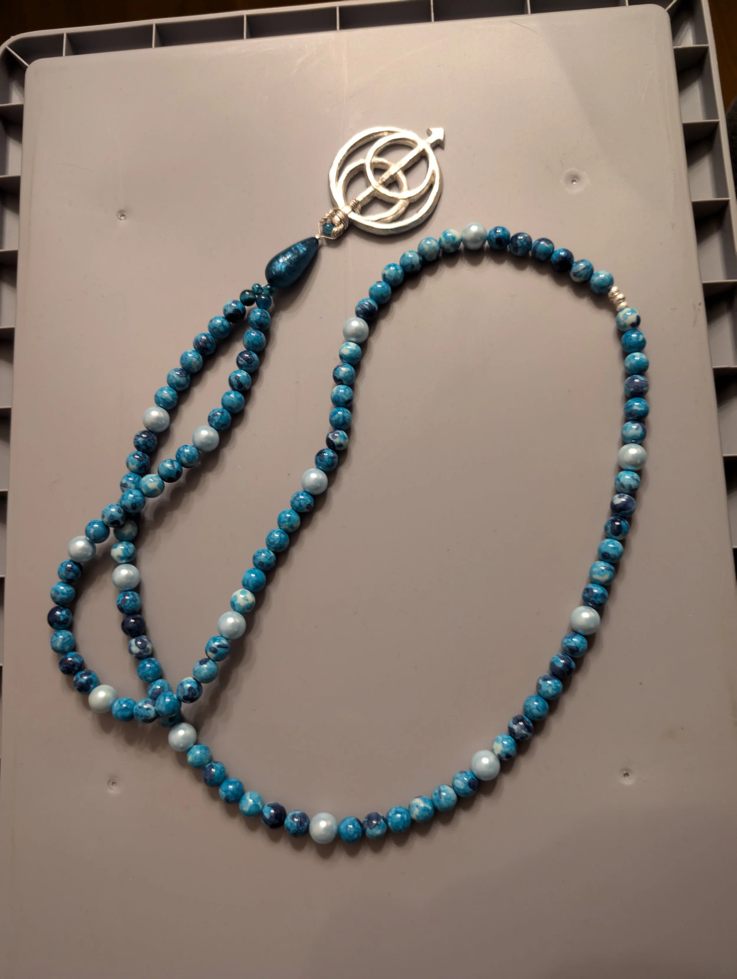 Halskette aus blauen und weißen Perlen mit einem silbernen Anhänger mit einem planetarischen Symbol.