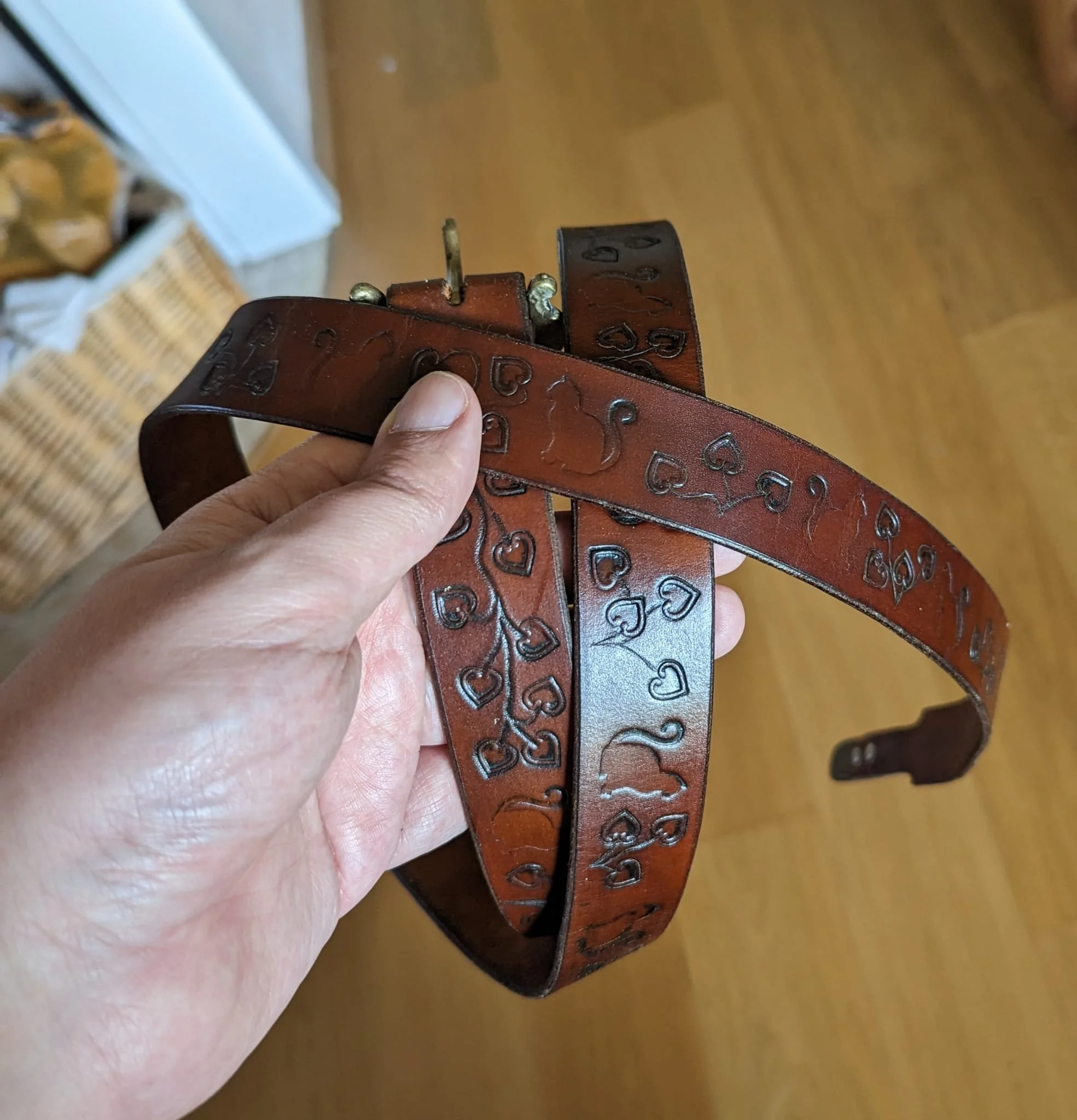 Brauner Ledergürtel mit eingravierten Herzen, gehalten in der Hand. Hintergrund zeigt Holzboden und ein Korb.