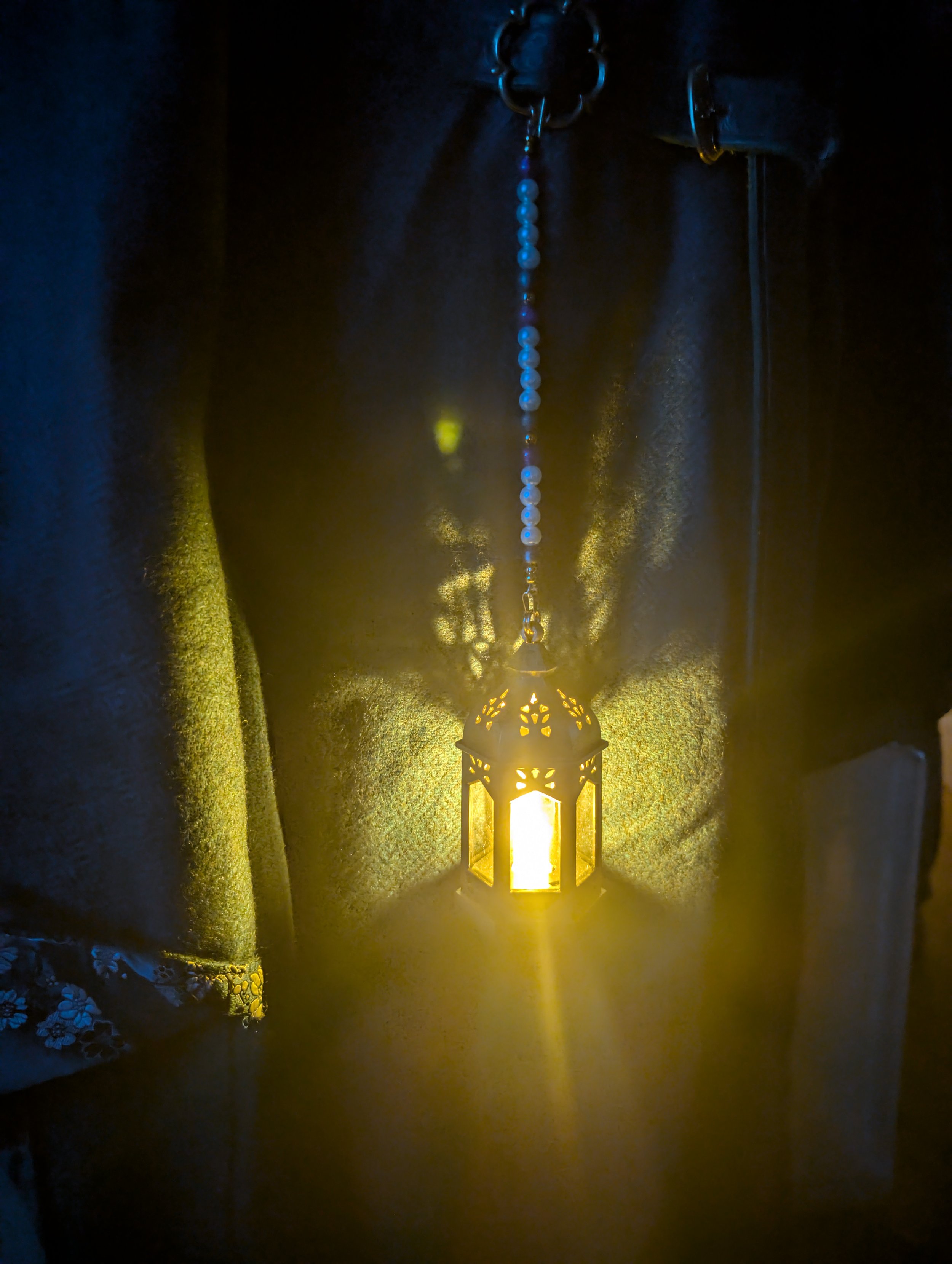 Ein gelber, orientalischer Laternenlampe, die an einer Kette von Perlen hängt, beleuchtet einen dunklen Raum und wirft Muster an die Wand.