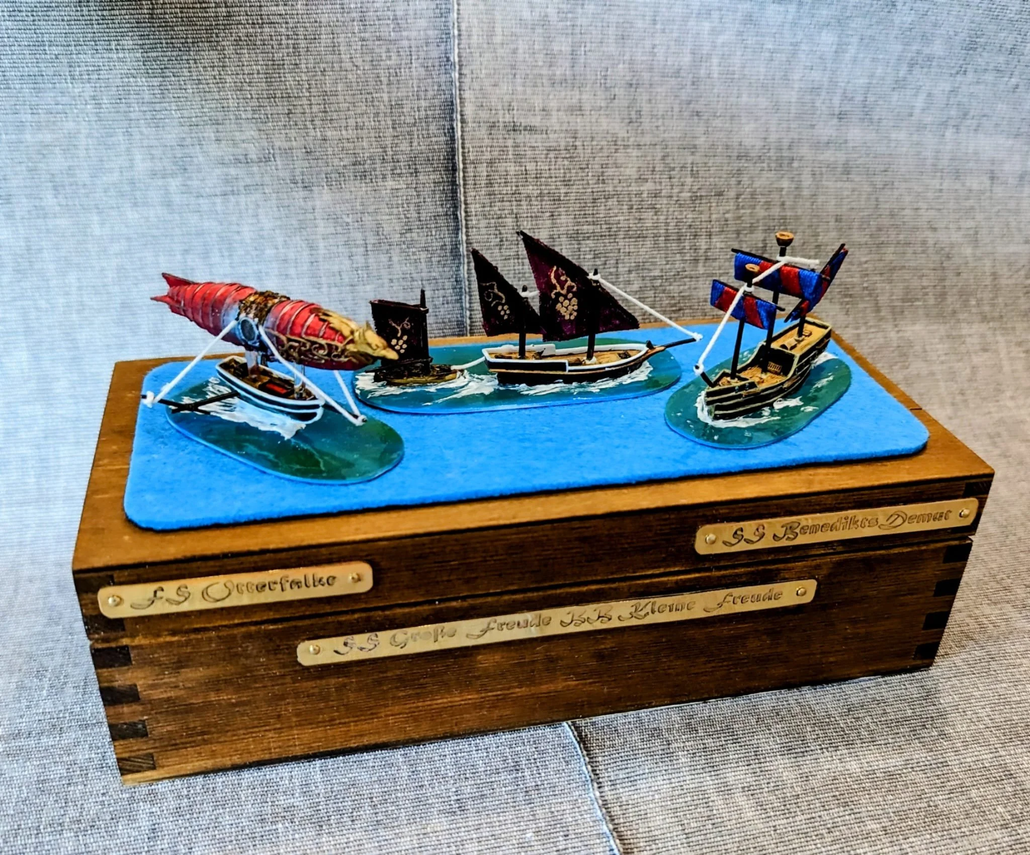 Eine Holzbox mit drei maritimen Spielzeugschiffen auf einer blauen Stoffunterlage.