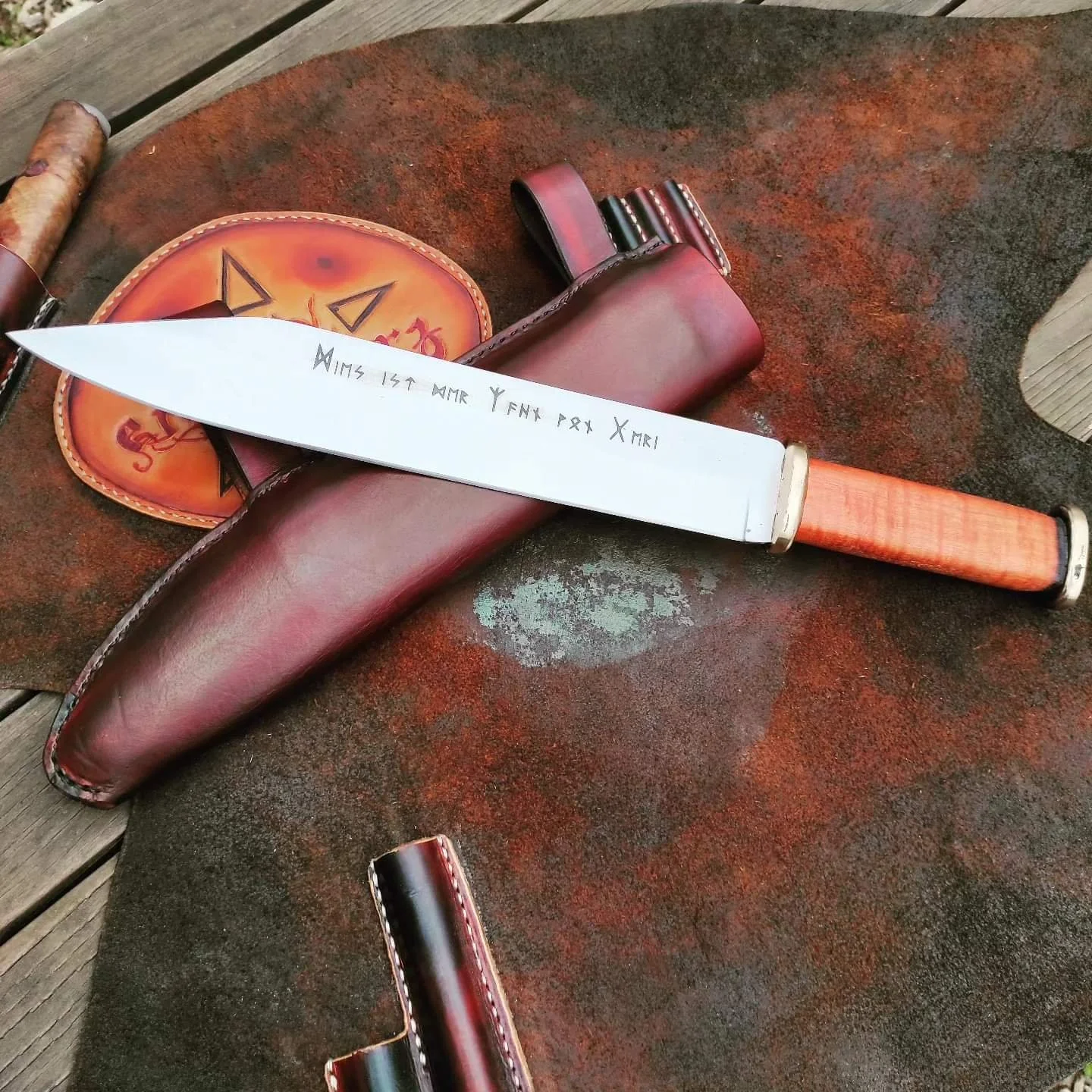 Ein Messer mit einer Klinge aus Metall, die eine Inschrift trägt, liegt auf einem rotbraunen Geschirrtuch. Das Messer hat einen Griff aus Holz. Im Hintergrund sind eine braune Lederscheide, ein orangefarbener Waschlappen mit einem Halloween-Gesicht u