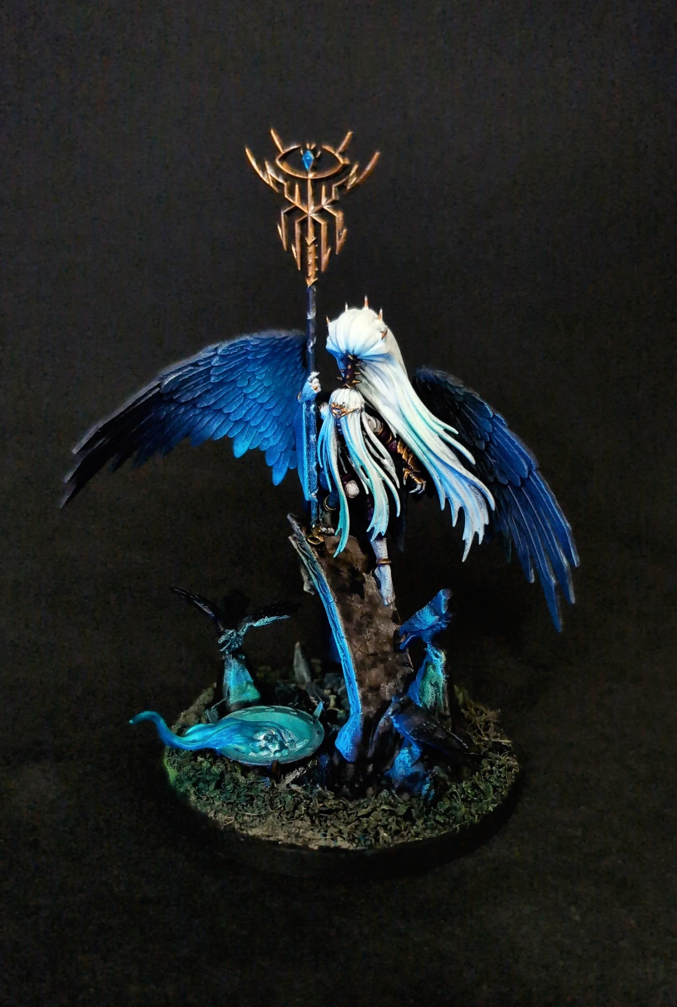 Detaillierte Miniaturfigur eines geflügelten Zauberers mit blauer und weißer Haare, der ein Sternstab hält, auf einer Basis mit Wasser-, Vogel- und Kryptidenähnlichen Figuren, vor schwarzem Hintergrund.