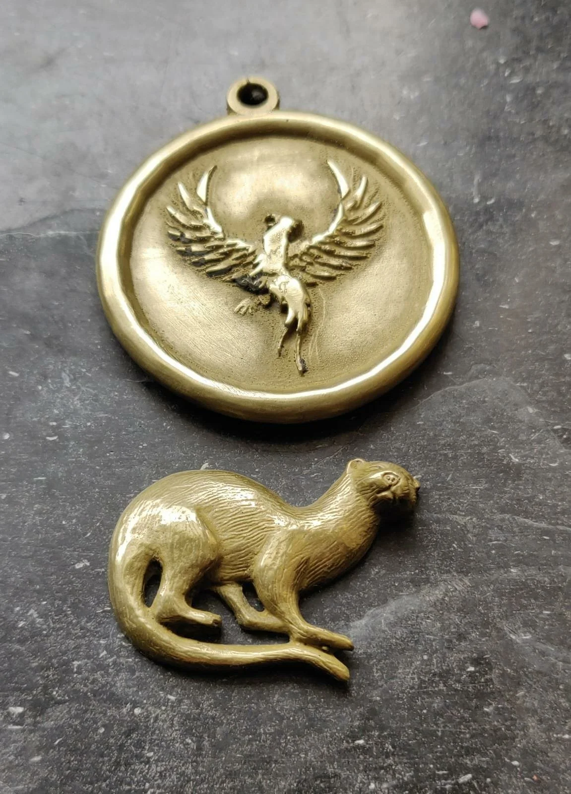 Bilder zeigen zwei goldene Schmuckstücke, ein Medaillon mit einem Phönix mit ausgebreiteten Flügeln und einem kleinen rundlichen Anhänger in Form einer sitzenden Hausratte.