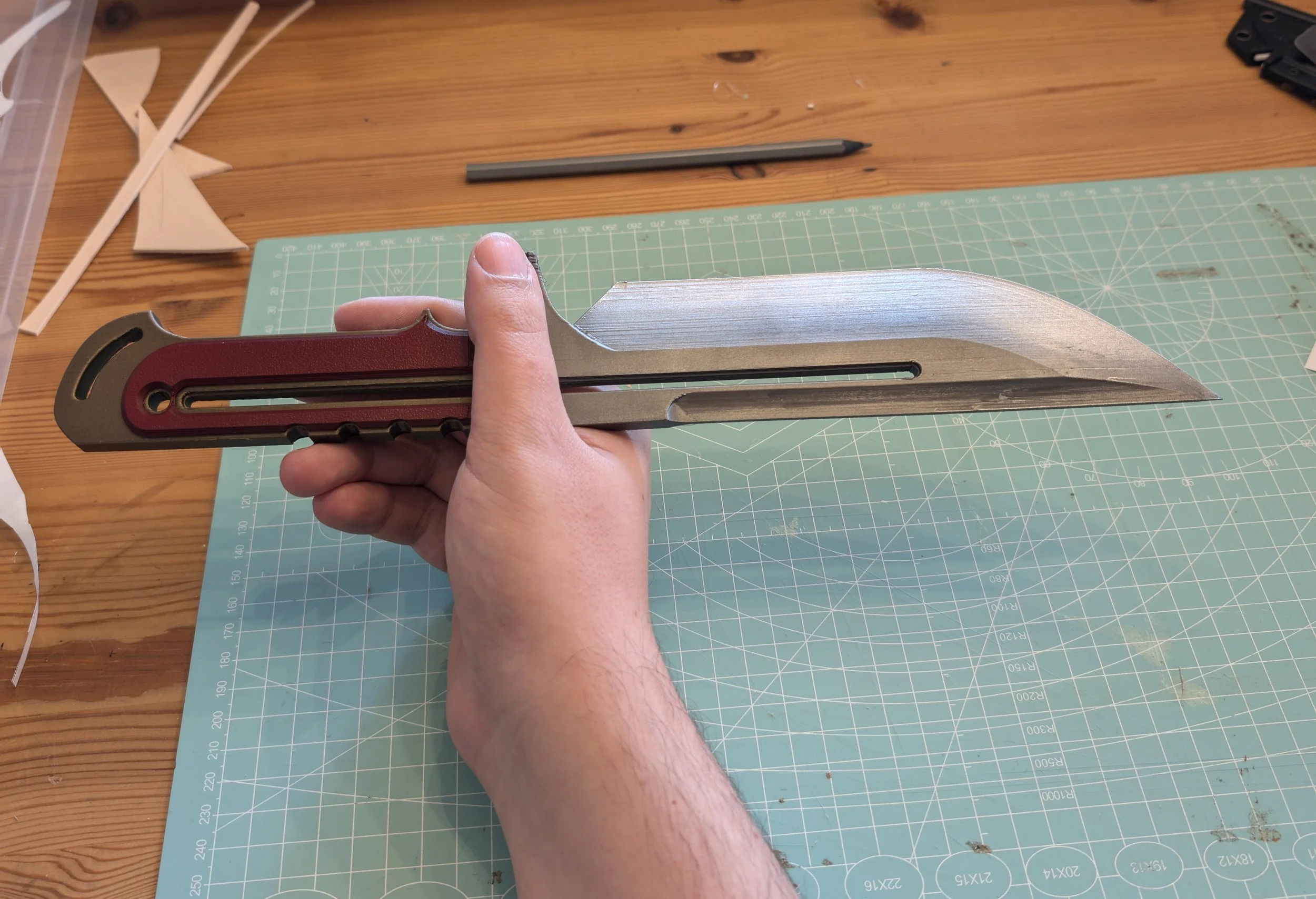 Eine Person hält ein großes, professionell aussehendes Kampfmesser oder Schwert mit einem langen, geschwungenen Blatt. Das Messer liegt über einem Schneidebrett aus Kunststoff auf einem Holztisch, umgeben von Bastelmaterialien.