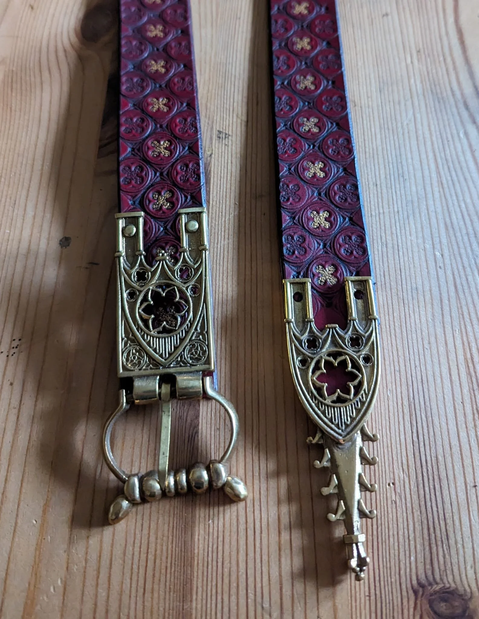 Zwei Gürtel mit roten Mustern und goldfarbenen Schnallen liegen auf einem Holztisch.