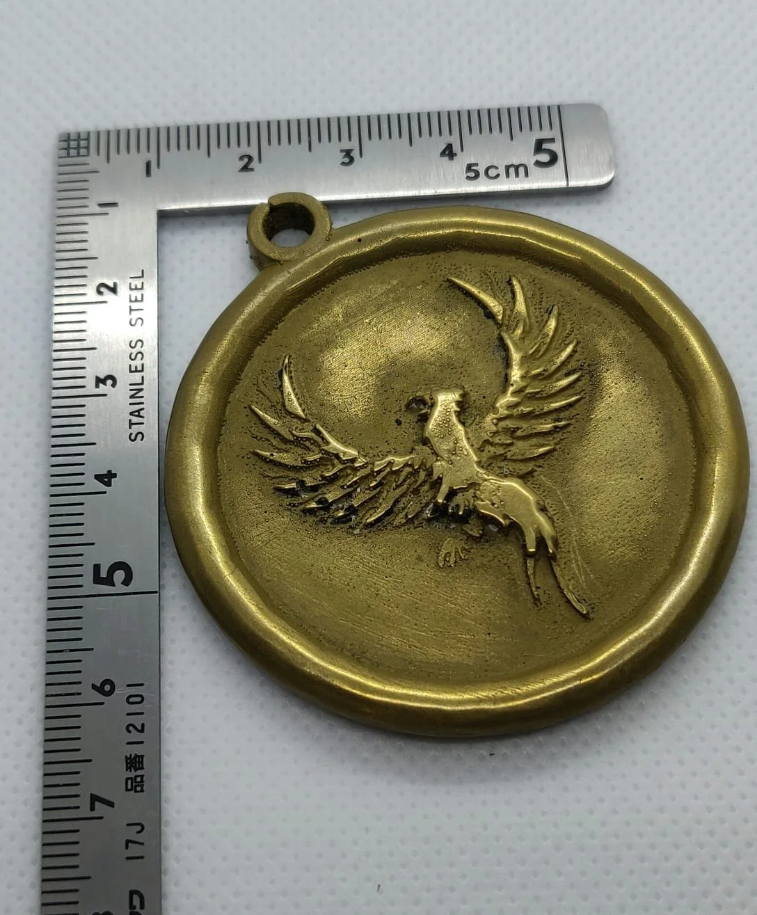 Eine goldene Medaille mit einem eingravierten Phönix in der Mitte, daneben ist ein Lineal, das die Medaille etwa 5 cm groß zeigt.