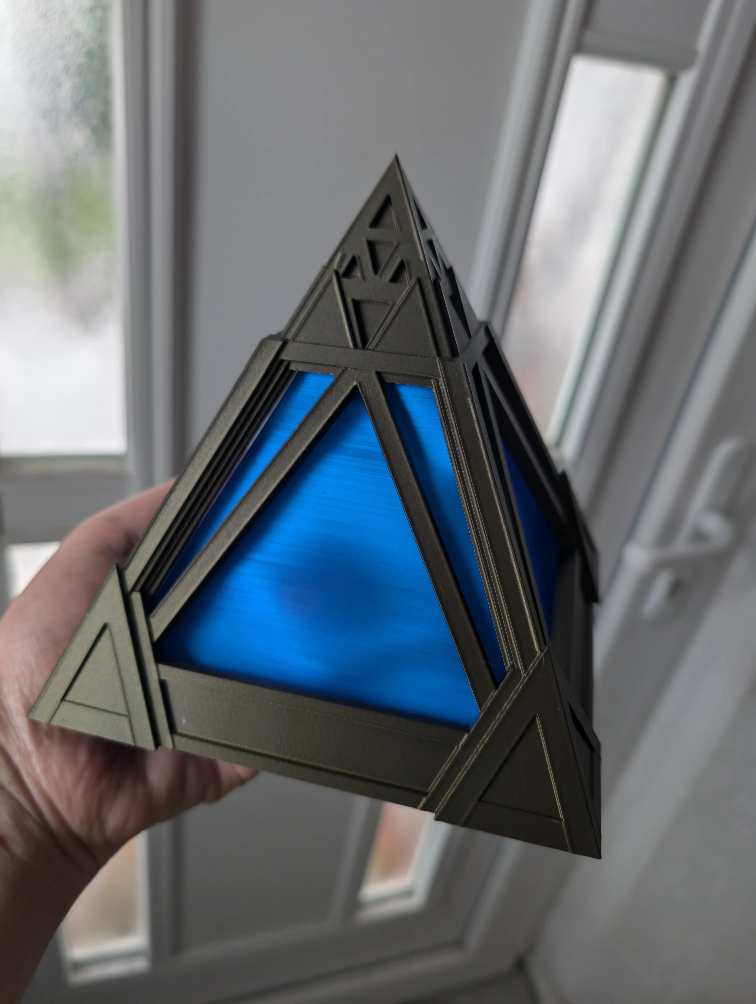 Eine Hand hält eine geometrische, pyramidenförmige Lampe mit blauer innerer Oberfläche vor einem Fenster mit weißen Rahmen.