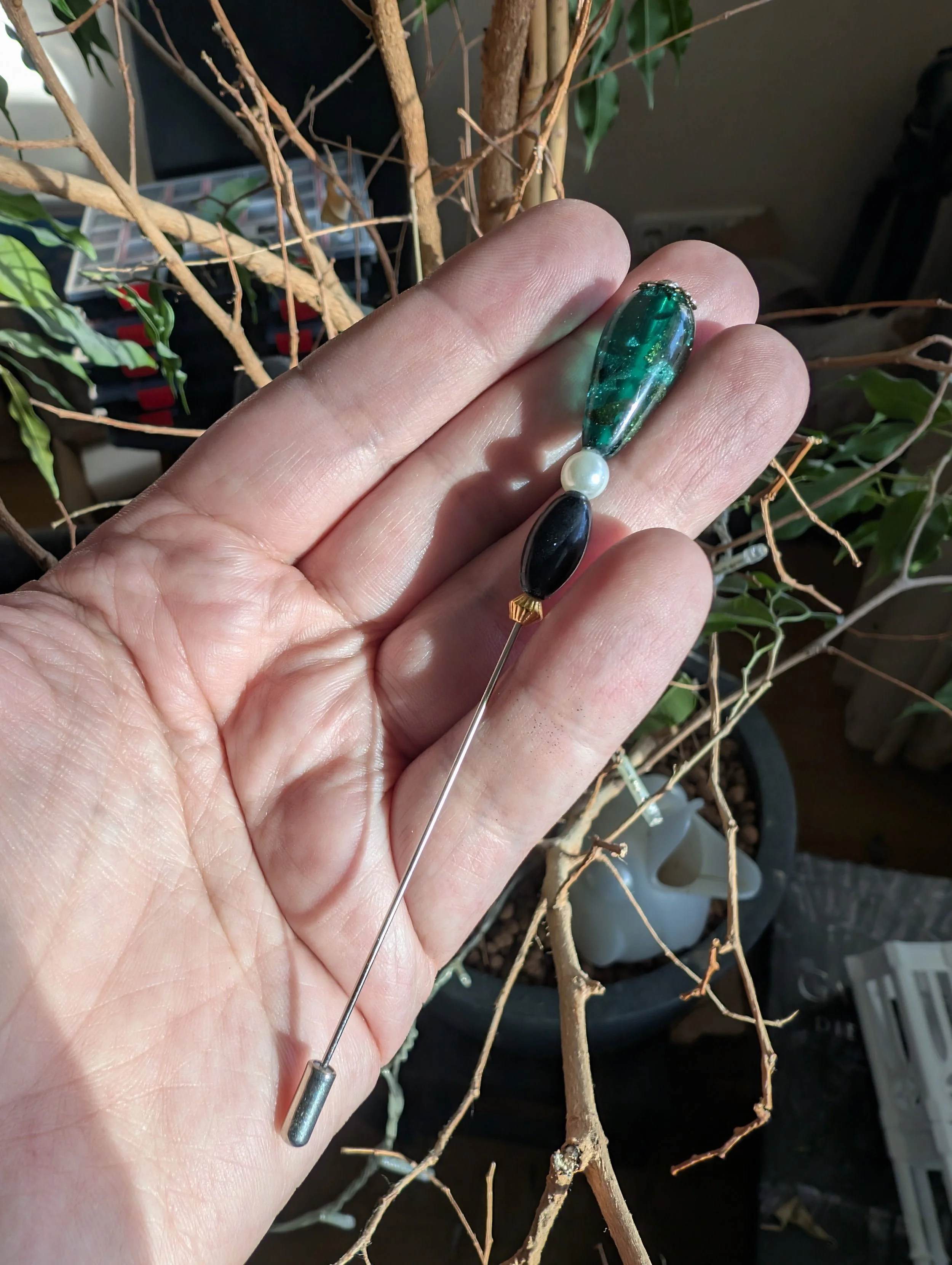 Nahaufnahme einer Hand, die eine dekorative Nadel mit mehreren Schmucksteinen hält, vor einem Topf mit einem Baum. Die Nadel hat einen langen, dünnen Metallstift mit einem kleinen Gewinde am Ende, darunter befinden sich verschiedenfarbige, glänzende 