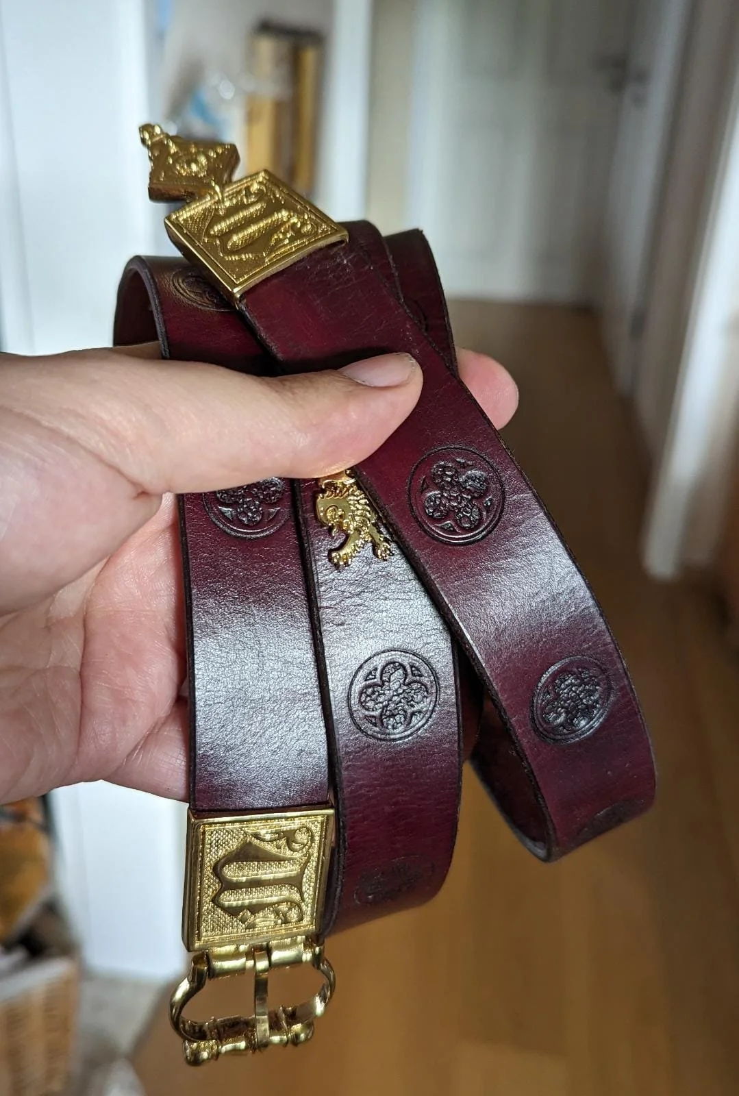 Hand hält einen braunen Ledergürtel mit goldenen Schnallen und dekorativen Verzierungen.