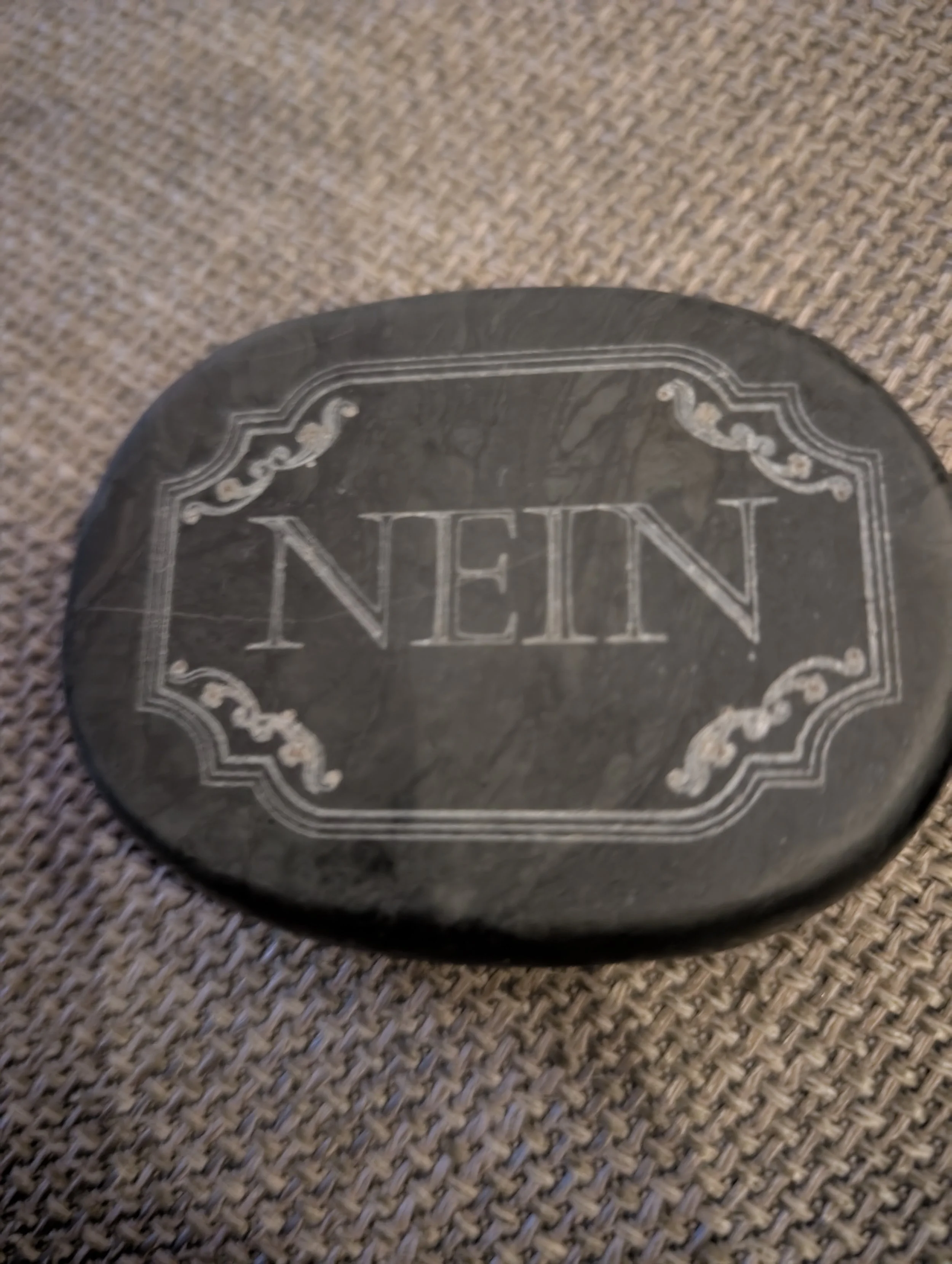 Runder dunkler Stein mit weißen gravierten Mustern und dem Wort 'NENE' in der Mitte, liegt auf einer braunen, gestrickten Oberfläche.