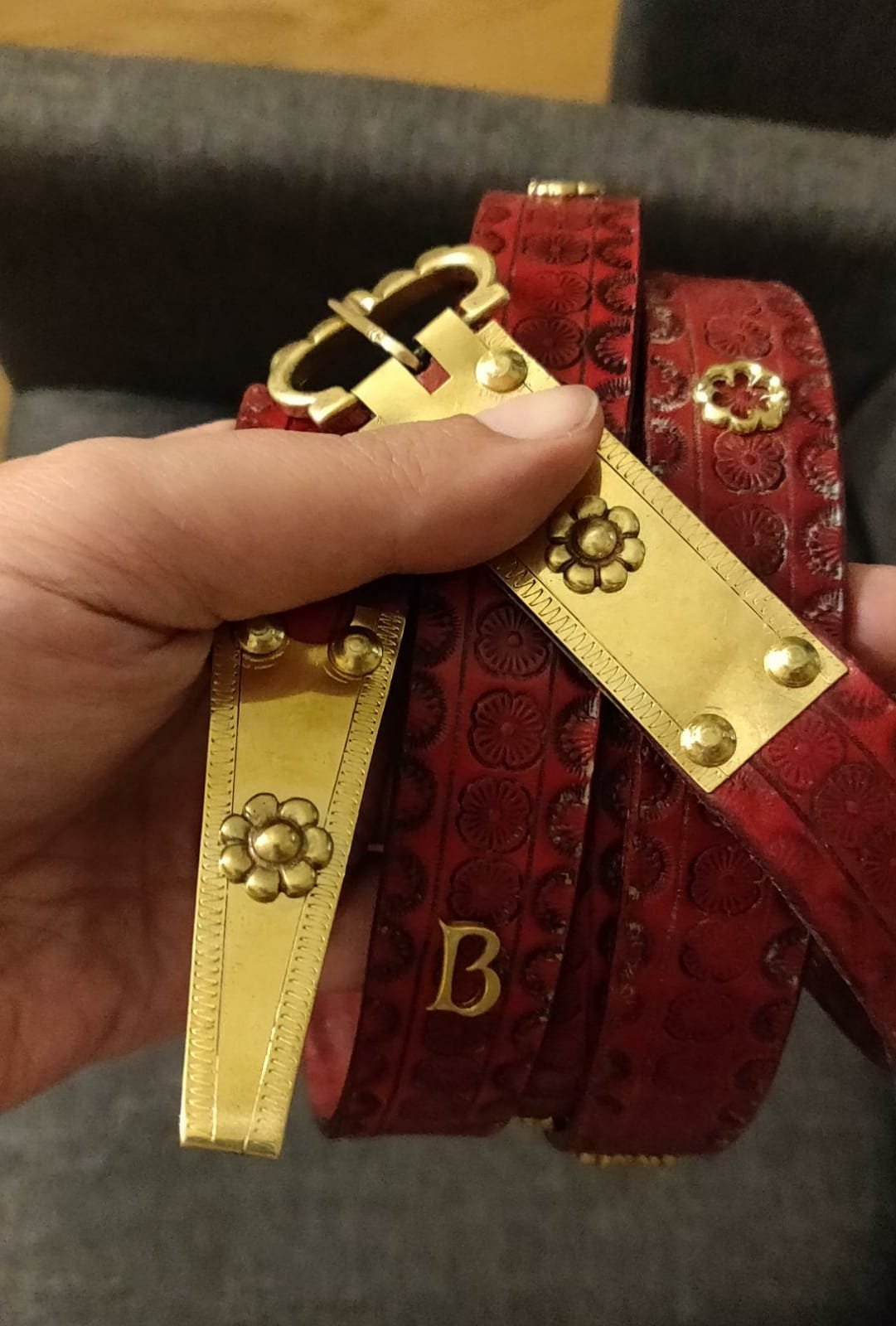 Hand hält einen goldenen Gürtel mit Blumenmuster und einem Buchstaben B, auf einem roten Hintergrund, mit Maßstab.