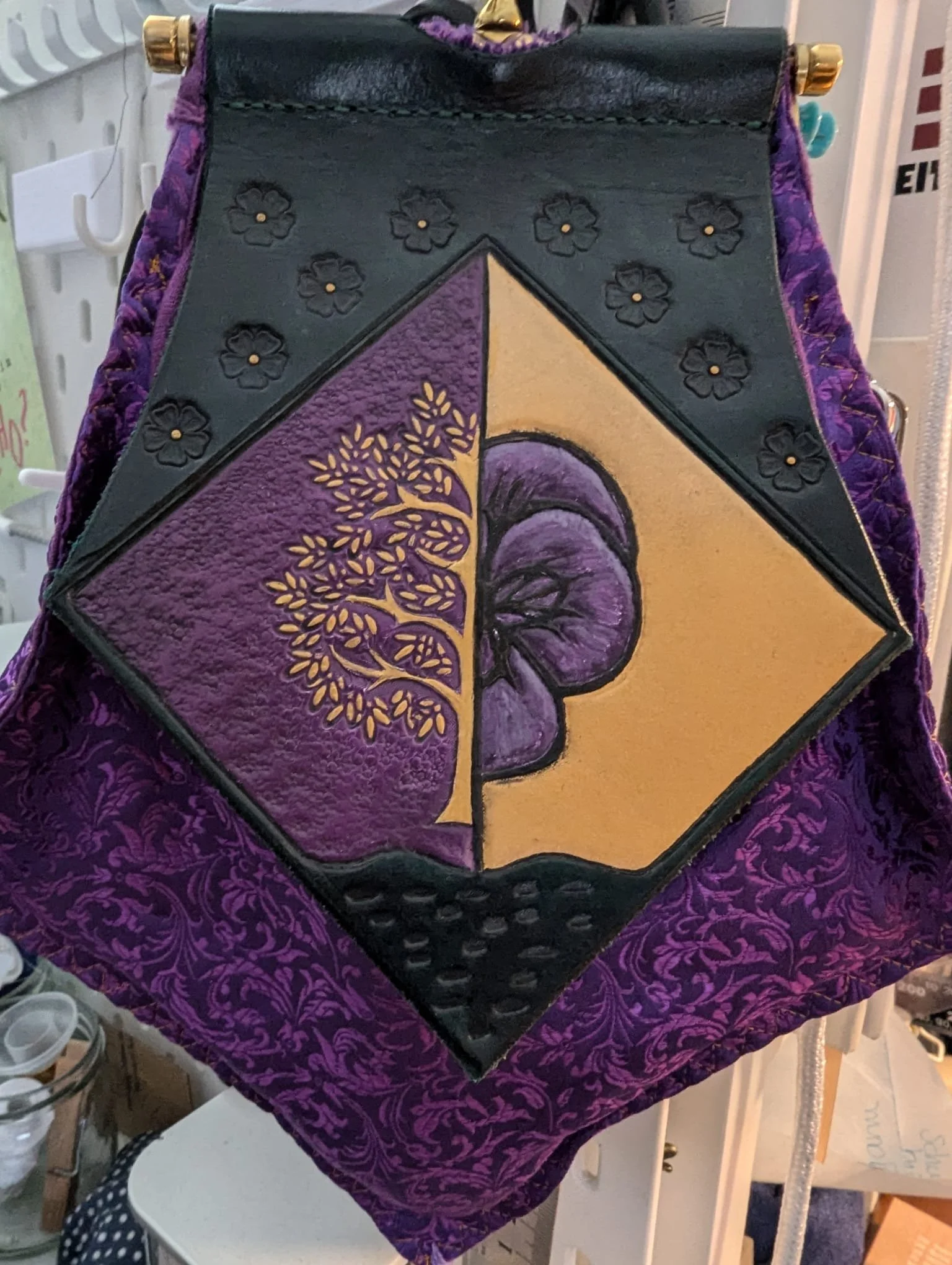 Handgemaltes Bild einer Pilz- und Baum-Szene, das in einem quadratischen Rahmen mit schwarzem Rand auf einem Stoff mit lila Muster befestigt ist.