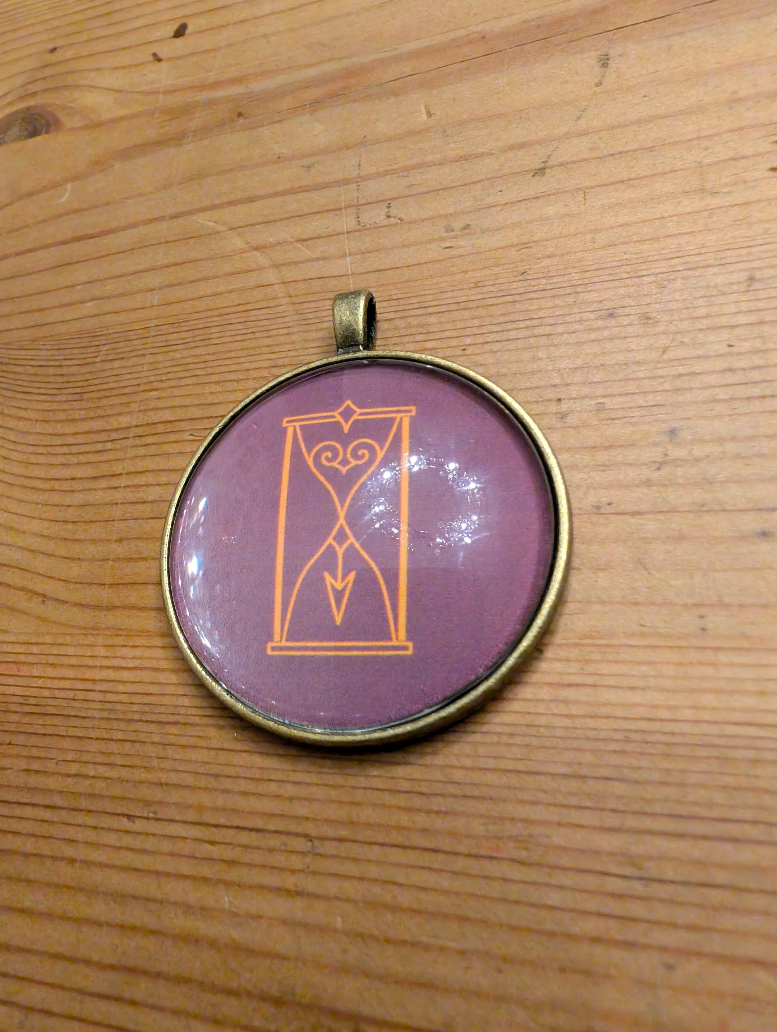 Runder Schmuckanhänger mit einem stilisierten, goldenen Linienmuster auf lila Hintergrund, auf Holzuntergrund liegend.