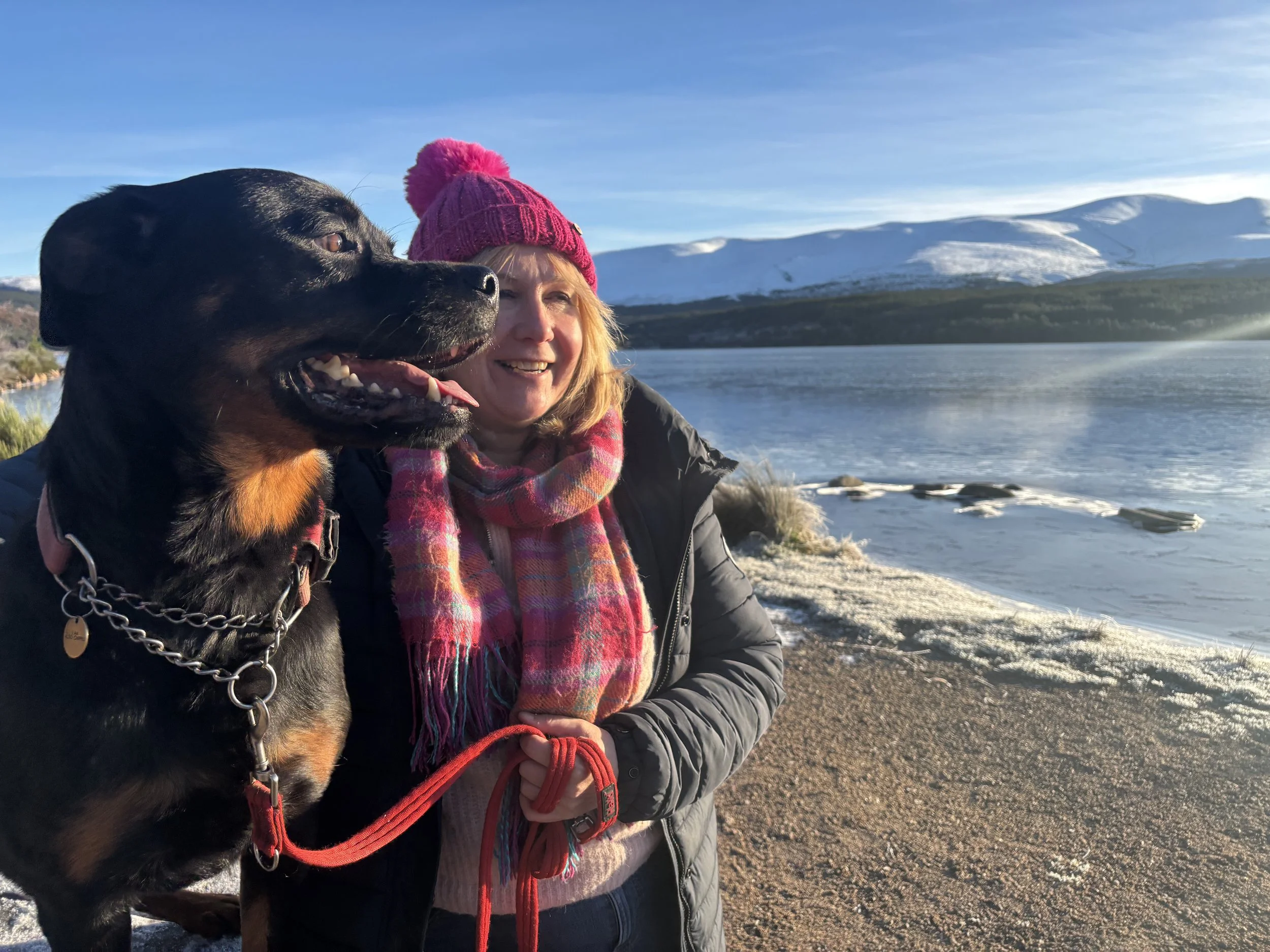 A woman in a pink hat and scarf standing next to a large black and tan dog near a lake with snow-covered mountains in the background.